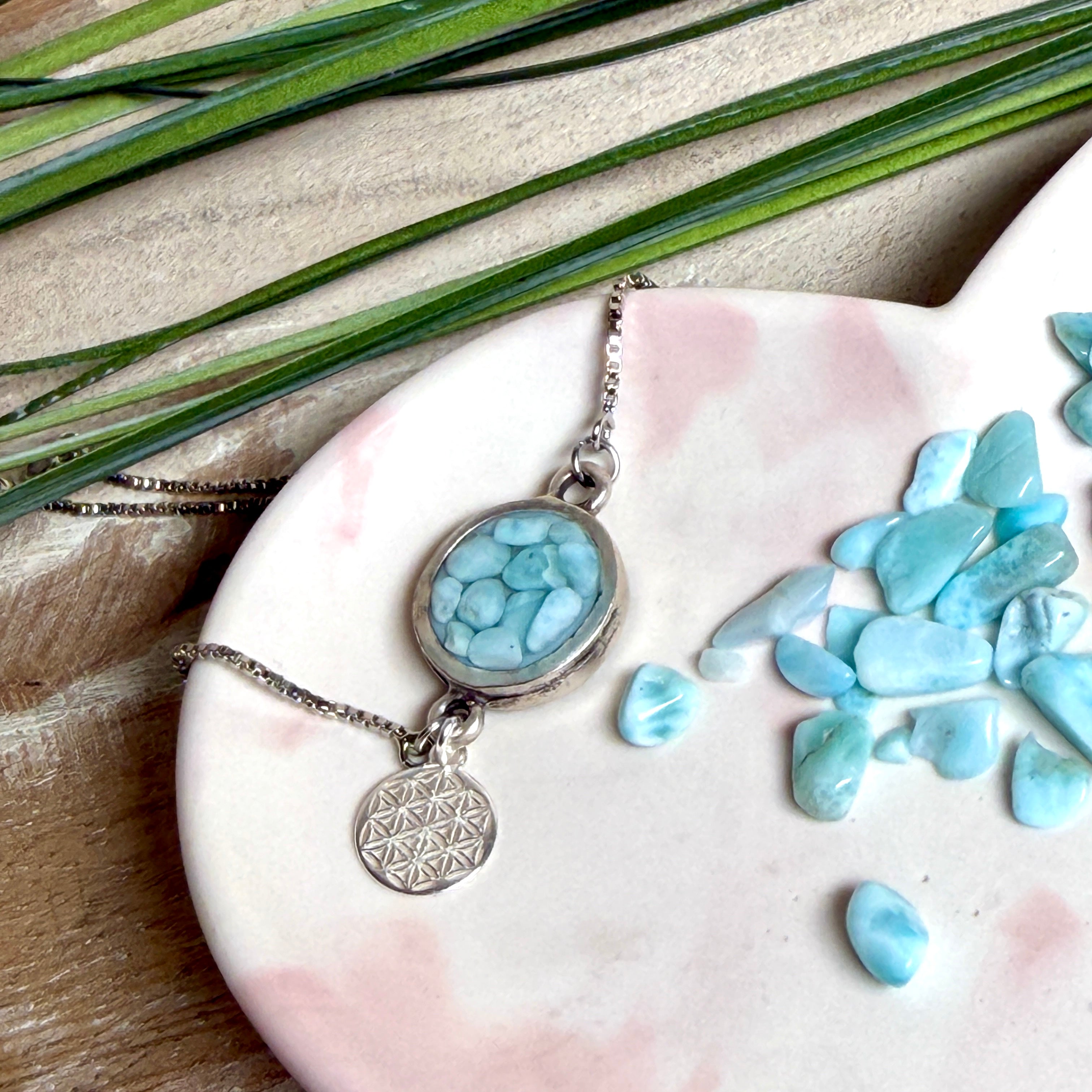 Verstellbares Edelstahlarmband mit Zirkoniasteinchen und Schiebeverschluss und rundem Anhänger (ca. 2,5 × 1,7 cm) mit echten Larimar-Edelstein-Splittern in Harz. Zusätzlich ein Charm „Blume des Lebens“ aus 925 Silber. Handgefertigtes Unikat in Türkis- und Meeresblautönen – Schmuckstück in Nahaufnahme bzw. als Tragebild am Handgelenk.