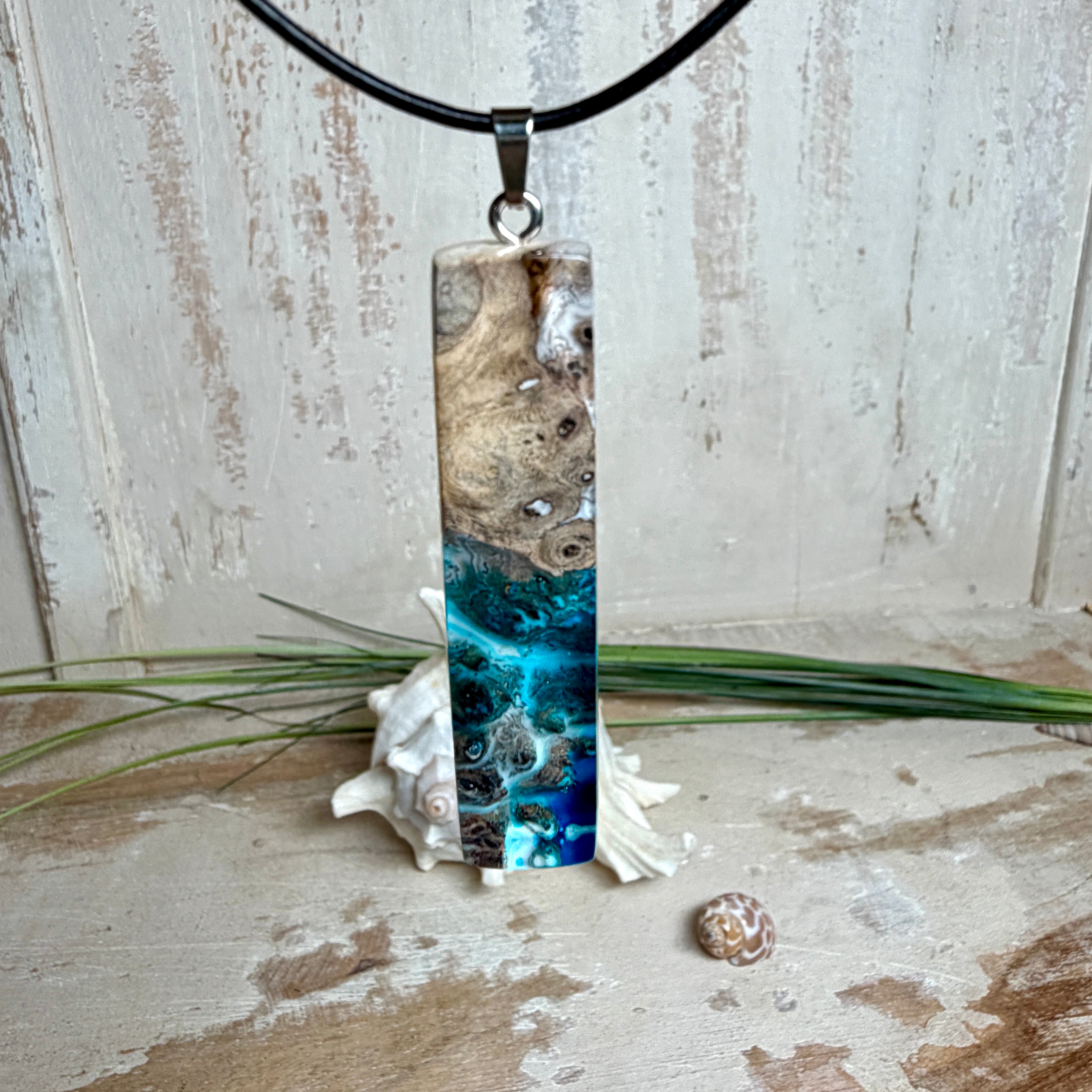 Rechteckiger Stabanhänger aus Maserholz und glasklarem Epoxidharz in tiefen Blau- und Türkistönen mit organischer Lagunen/Riff-Optik. Hochglanzpoliert, ca. 5,3 × 1,2 cm. Der Anhänger hängt an einer schwarzen Lederkette mit Edelstahl-Karabiner und 5-cm-Verlängerung (45 + 5 cm); die Anhängerschlaufe ist ebenfalls aus Edelstahl. Handgefertigtes Unikat, Lieferung in edler Schmuckbox – maritimer Look.