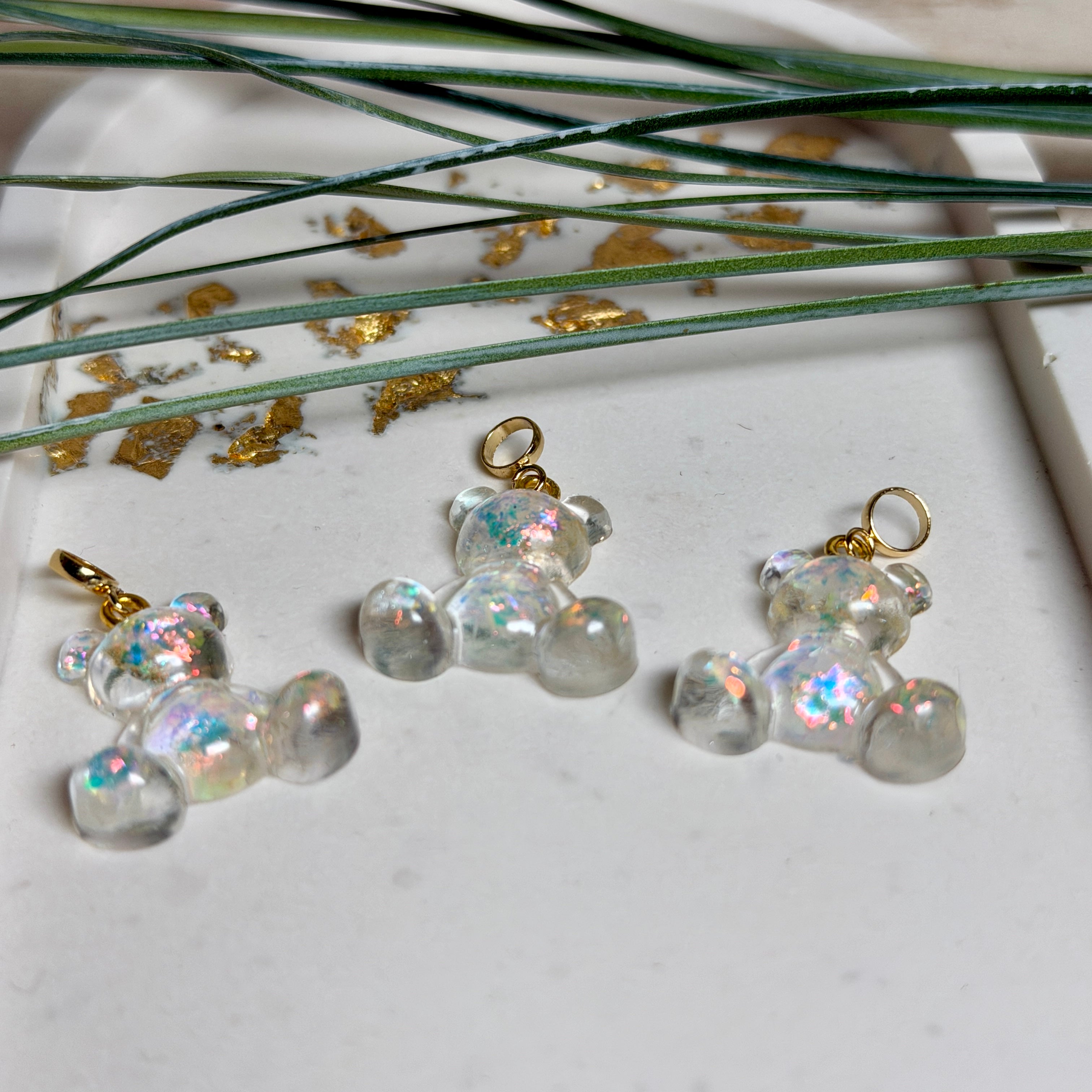 Kleiner Bärchen-Anhänger aus transparentem Harz mit irisierenden Flakes in Regenbogen-Schimmer, goldfarbene Anhängerschlaufe aus Edelstahl. Größe ca. 2,2 × 2,1 cm. Handgefertigtes Unikat; Flakes-Anordnung variiert, vereinzelt kleine Luftbläschen möglich.