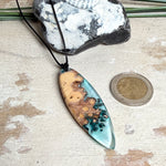 Handgefertigter, länglicher Surfbrett-Anhänger aus naturbelassenem Maserholz und transparentem Harz in Lagunen-Türkis, an schwarzem Lederband mit Knoten – maritimer Unikatschmuck für Damen und Herren, dekorativ mit Stein und Muschel inszeniert.