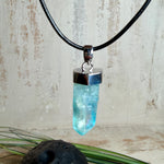Nahaufnahme einer handgefertigten Halskette mit Aqua Aura Rohkristall: naturbelassener, sechskantiger Bergkristall mit goldbedampfter Aqua-Beschichtung in Türkisblau, gefasst in 925er Silber und getragen an einer schwarzen Lederkette mit Edelstahl-Karabiner und Verlängerungskettchen.