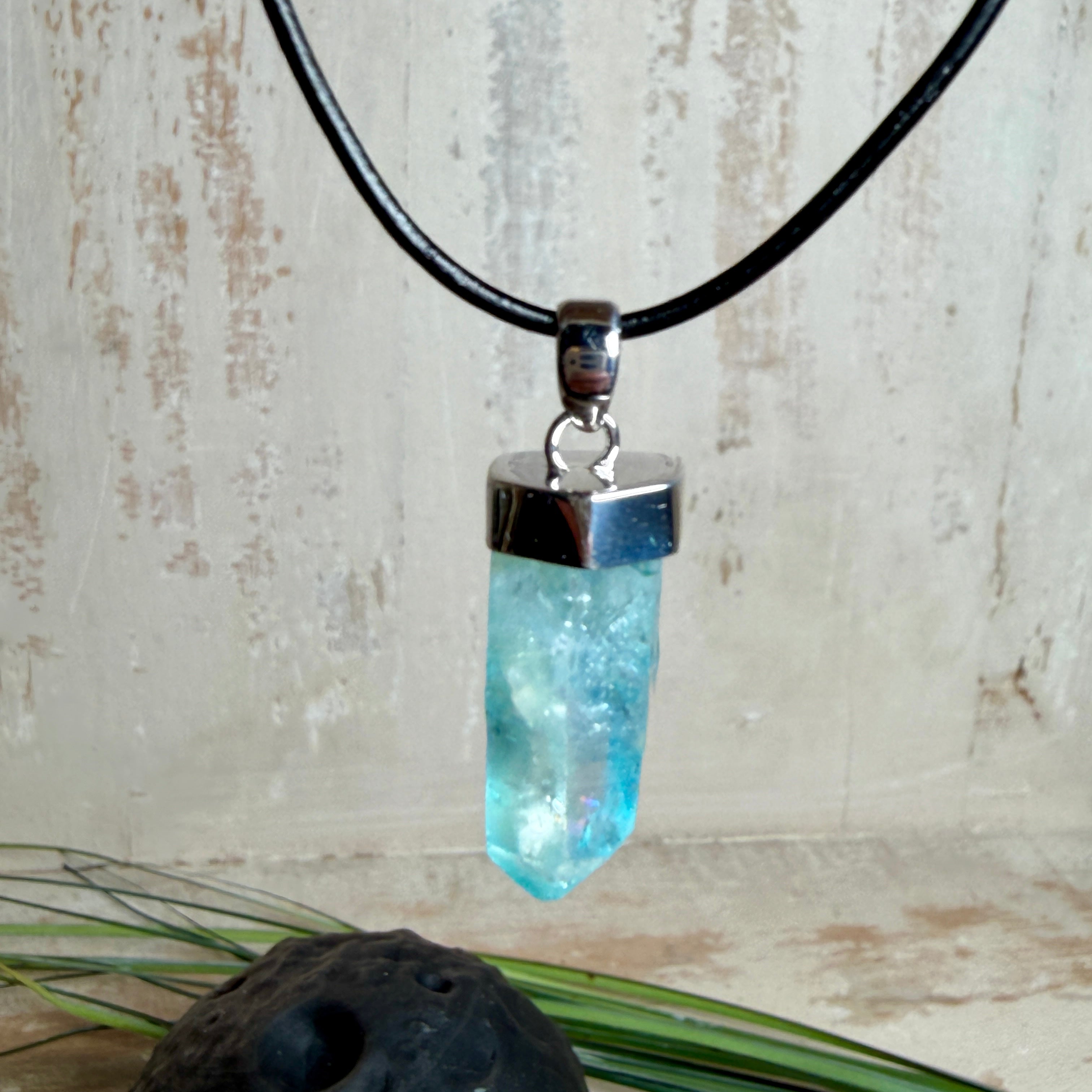 Nahaufnahme einer handgefertigten Halskette mit Aqua Aura Rohkristall: naturbelassener, sechskantiger Bergkristall mit goldbedampfter Aqua-Beschichtung in Türkisblau, gefasst in 925er Silber und getragen an einer schwarzen Lederkette mit Edelstahl-Karabiner und Verlängerungskettchen.