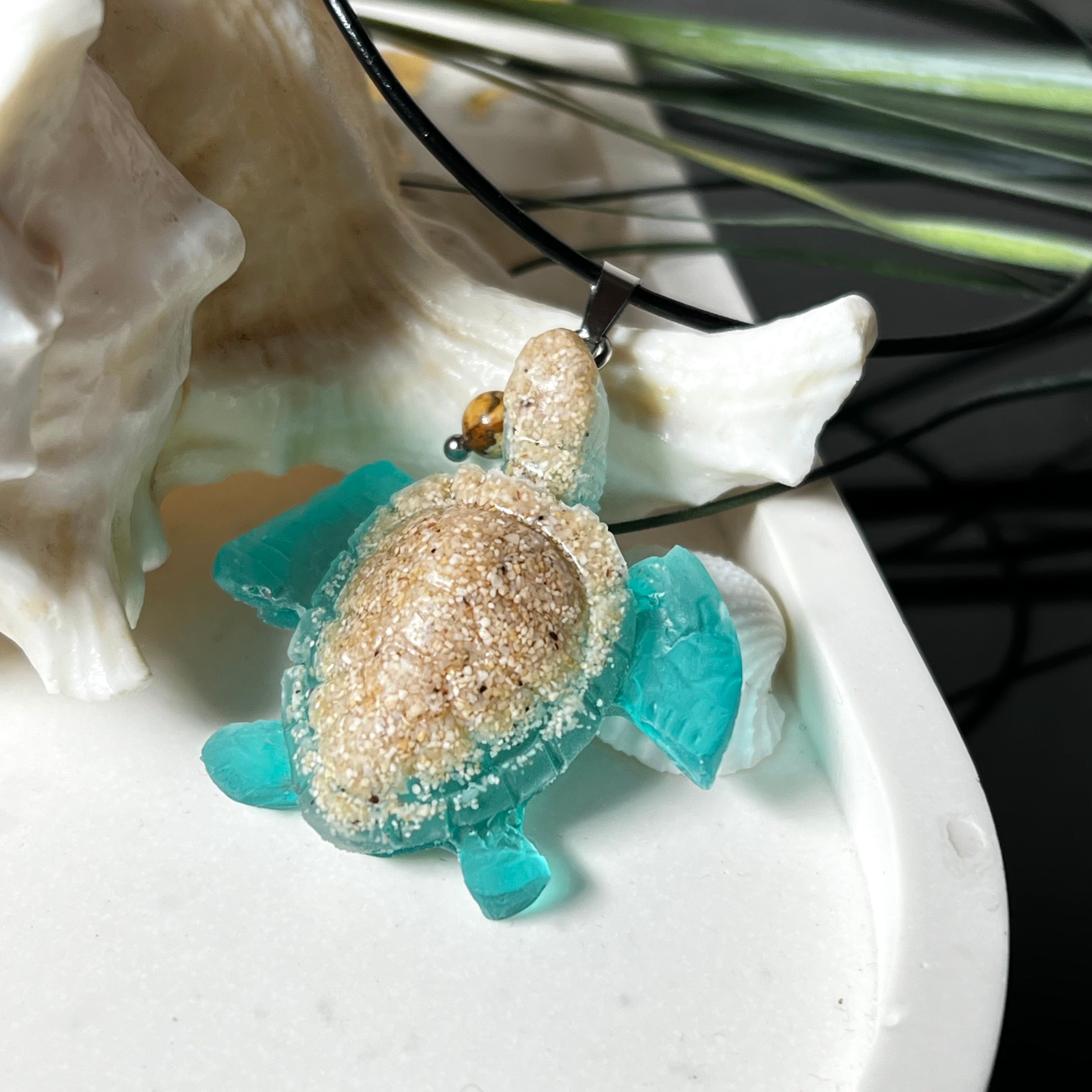 Handgefertigter Schildkröten-Anhänger Turtle Harmony aus echtem Meersand und transparent-türkisfarbenem Harz, kombiniert mit einer kleinen Jaspisperle. Symbol für Weisheit, Schutz und Harmonie. Mit Edelstahl-Anhängerschlaufe und Lederkette (45 + 5 cm). Maritimer Schmuck als Unikat, ideal als liebevolles Geschenk oder stilvolles Accessoire für den Alltag.