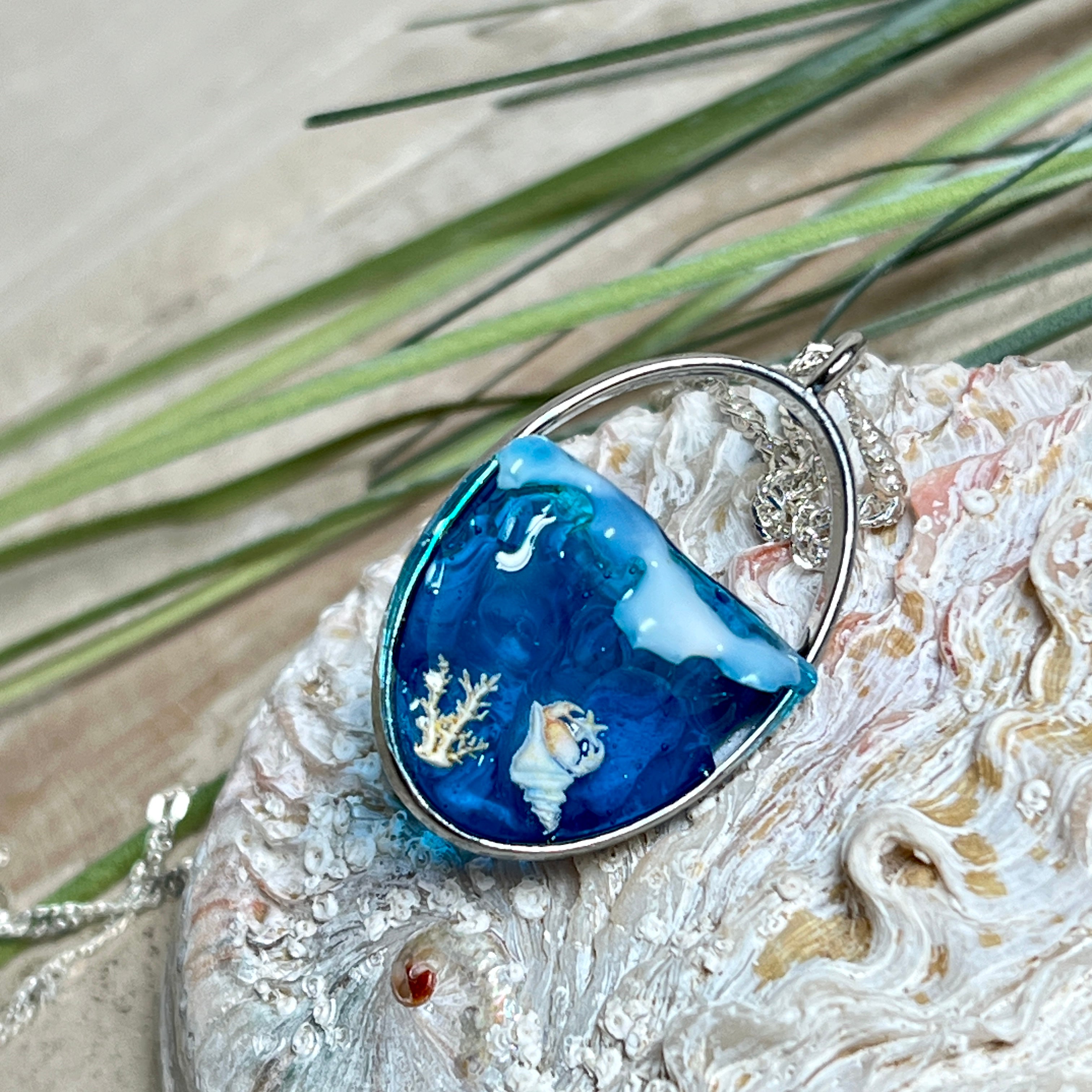 „Riffschatz“ – ovaler Harz-Anhänger mit plastischer 3D-Welle in Blau/Türkis, weißer Schaumkrone und Muschel-/Korallen-Stickern am Grund. Die Sticker sind unter klarem Harz versiegelt. Silber-Legierung, ca. 3,9 × 2,3 cm, an einer ca. 56 cm langen versilberten Kette. Handgefertigt, hochglänzend, jedes Stück ein Unikat.