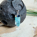 Nahaufnahme einer handgefertigten Halskette mit Aqua Aura Rohkristall: naturbelassener, sechskantiger Bergkristall mit goldbedampfter Aqua-Beschichtung in Türkisblau, gefasst in 925er Silber und getragen an einer schwarzen Lederkette mit Edelstahl-Karabiner und Verlängerungskettchen.