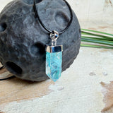 Nahaufnahme einer handgefertigten Halskette mit Aqua Aura Rohkristall: naturbelassener, sechskantiger Bergkristall mit goldbedampfter Aqua-Beschichtung in Türkisblau, gefasst in 925er Silber und getragen an einer schwarzen Lederkette mit Edelstahl-Karabiner und Verlängerungskettchen.