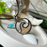 Wave Spirit – runder Edelstahlanhänger mit echter Sandfüllung in Harz eingebettet, inspiriert von einer stilisierten Welle. Verziert mit zwei blauen Achatperlen. Der Anhänger (ca. 2,5 cm Durchmesser) hängt an einer 45 cm langen Lederkette mit Edelstahlverschluss und 5 cm Verlängerungskette. Maritimer Look trifft moderne Eleganz – ein handgefertigtes Schmuckstück für Meerliebhaber
