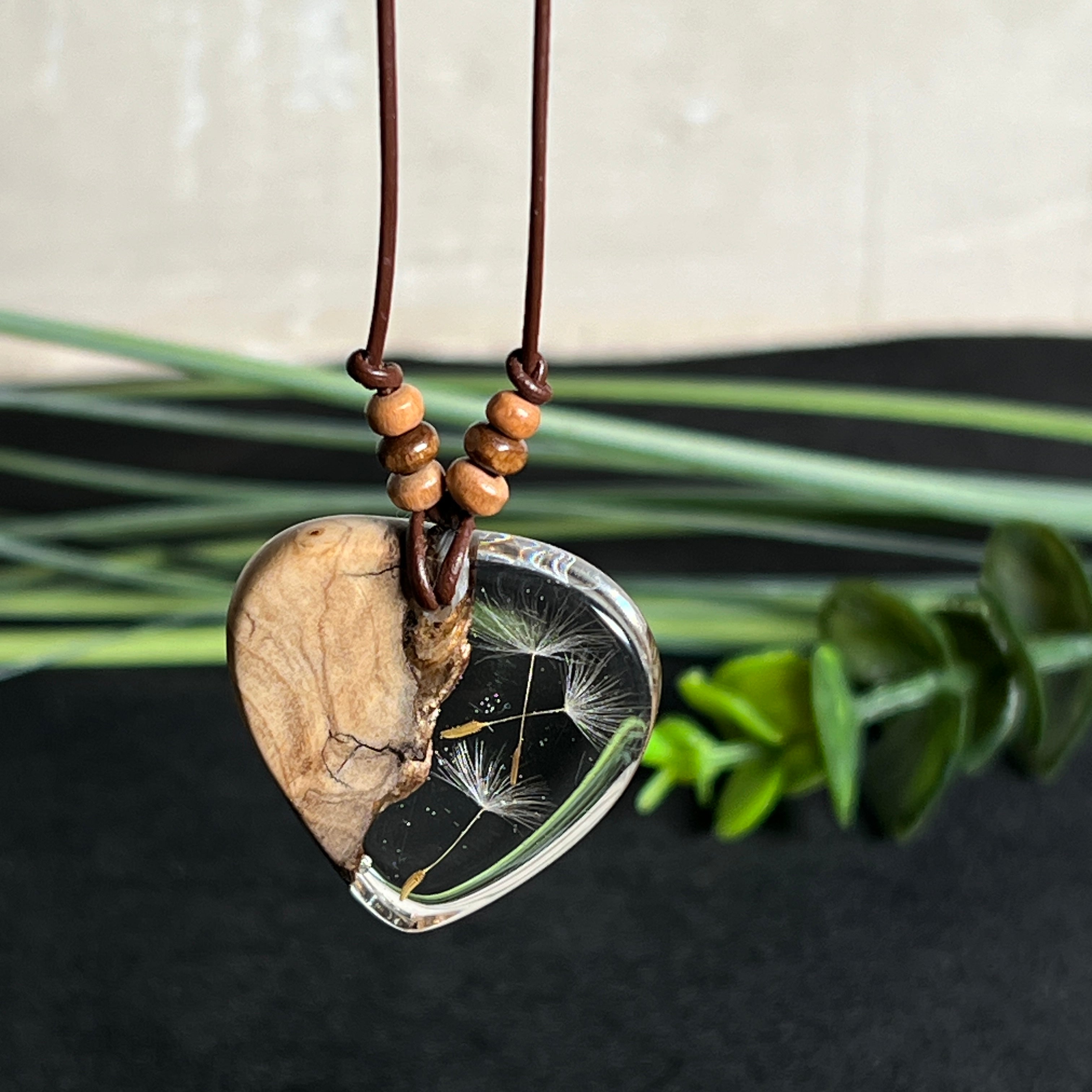 Handgefertigter Herzanhänger aus Olivenholz und klarem Harz mit echten Pusteblumen. Der Anhänger misst ca. 3,5 cm, das Lederband ist verstellbar von 23–43 cm und mit kleinen Holzperlen verziert. Ein natürliches Unikat, Symbol für Wünsche und Glück – sorgfältig gegossen, gesägt, geschliffen und poliert.