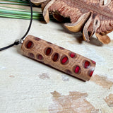 Handgefertigter Halsanhänger aus echtem Banksiazapfen, veredelt mit rotem Harz in den natürlichen Kammern; ca. 5,5 × 1,6 cm. Mit Anhängerschlaufe aus Edelstahl an schwarzer Lederkette (45 cm + 5 cm Verlängerung) mit Edelstahl-Karabiner. Detailaufnahme bzw. Tragebild vor neutralem, natürlichem Hintergrund.