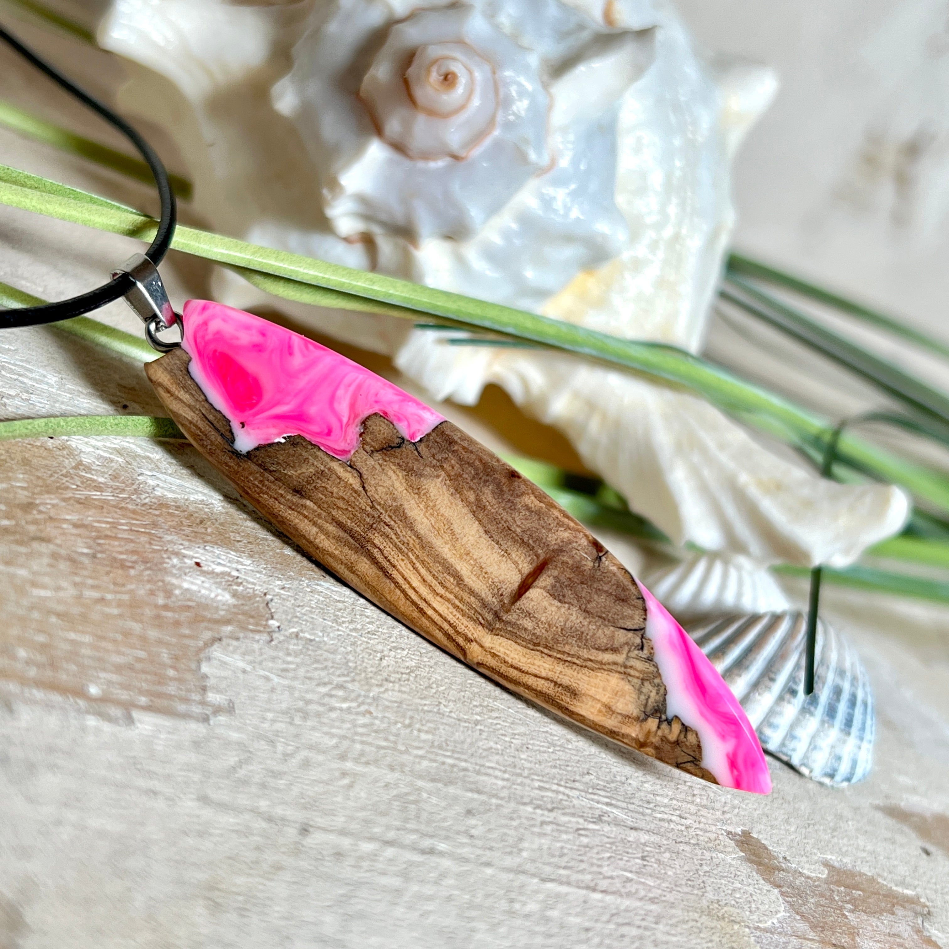 Handgefertigter Surfbrett-Anhänger „Pink Glow“ aus edlem Olivenholz und fluoreszierendem Epoxidharz in Pink & Weiß. Fluoreszierend bei Schwarzlicht, mit Edelstahl-Anhängerschlaufe und schwarzer Lederkette (45 cm + 5 cm Verlängerung). Ein maritimes Unikat – perfekt als Geschenk für Surfer und Meeresliebhaber.