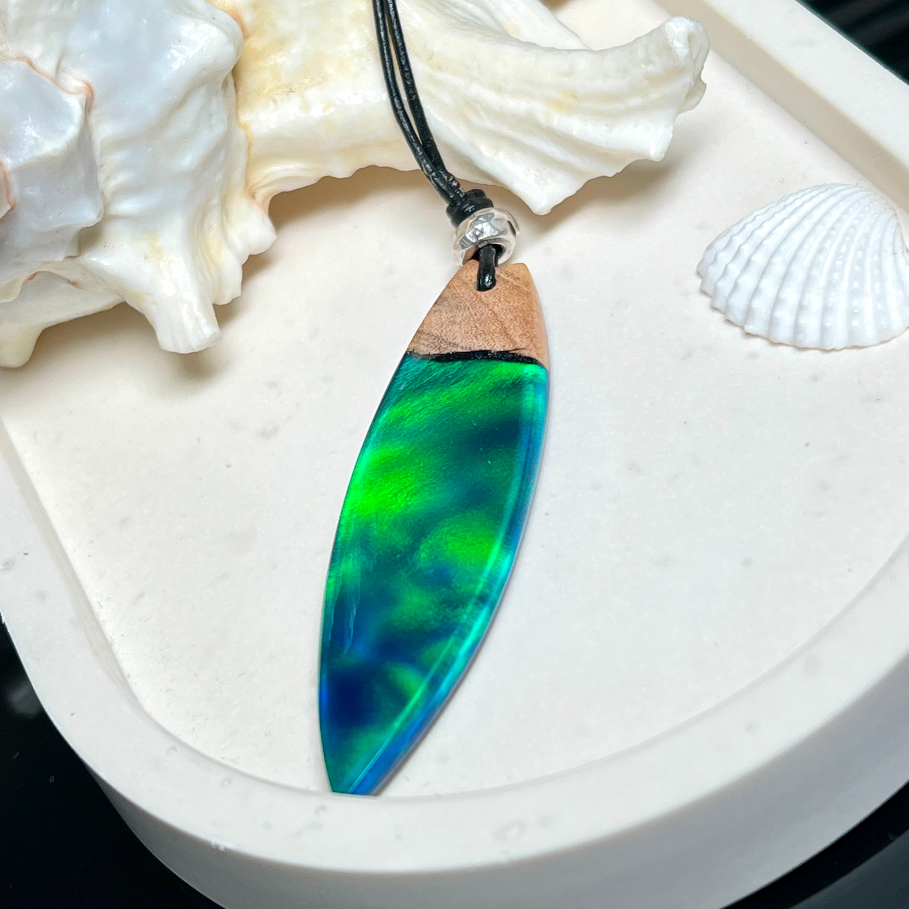 Beidseitig tragbarer Surfbrett-Anhänger aus Olivenholz, kombiniert mit klaren Epoxidharz in Blau-Grün. Eingearbeiteter synthetischer Opal erzeugt ein faszinierendes Farbspiel wie Nordlichter. Größe ca. 6 × 1,8 cm. Mit verstellbarem Lederband (26–45 cm), dekorativen Perlen & Schiebeknoten. Handgefertigt & einzigartig. Ein Schmuckstück mit Naturinspiration.