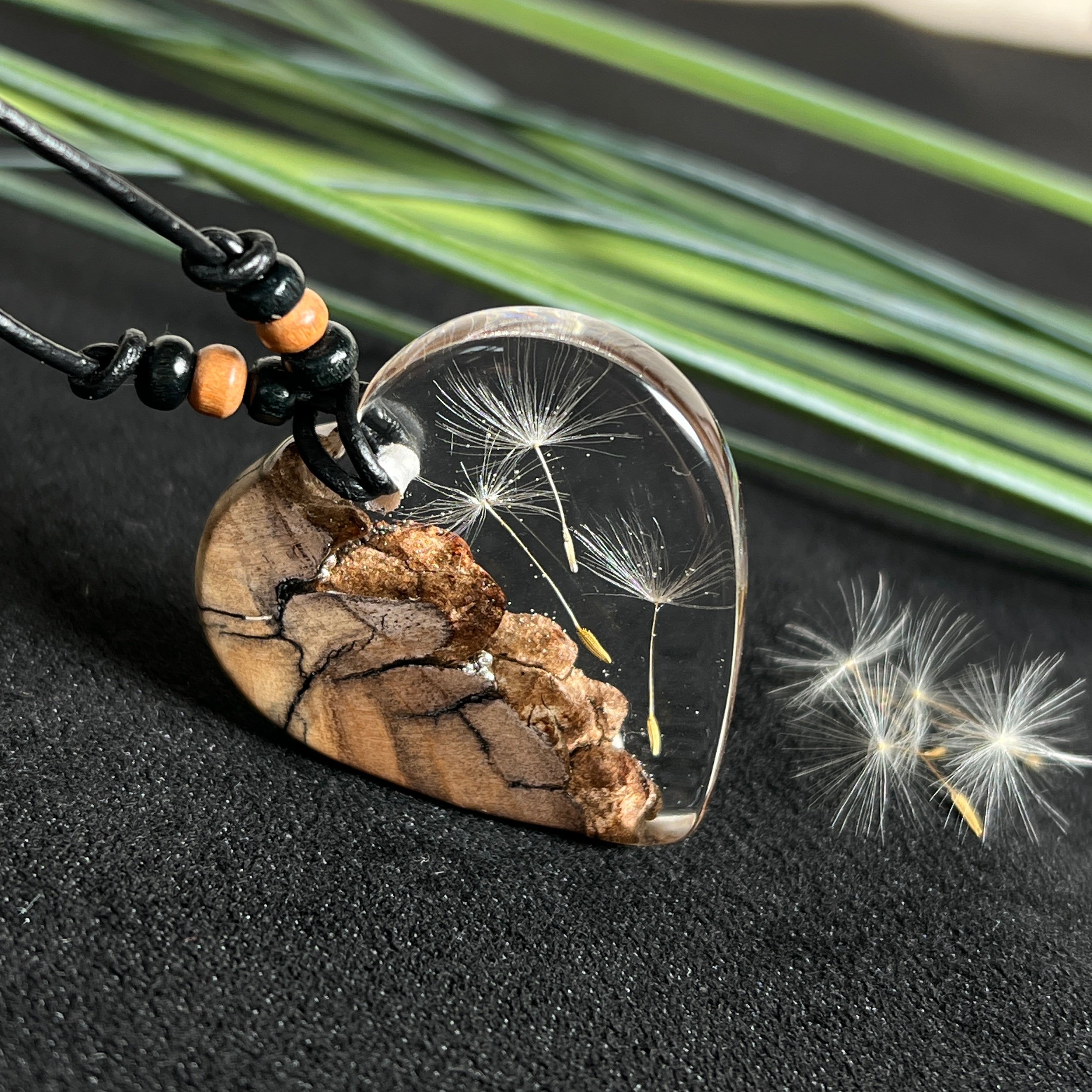 „Handgefertigter Herzanhänger aus Olivenholz und klarem Harz mit echter Pusteblume. Der Anhänger (ca. 3,5 × 3,5 cm) hängt an einem verstellbaren Lederband (23–43 cm) mit Holzperlen. Symbol für Wünsche und Glück, sorgfältig in Handarbeit gegossen, gesägt, geschliffen und poliert. Ein einzigartiger, naturinspirierter Anhänger – ideal als Geschenk oder persönlicher Talisman.“