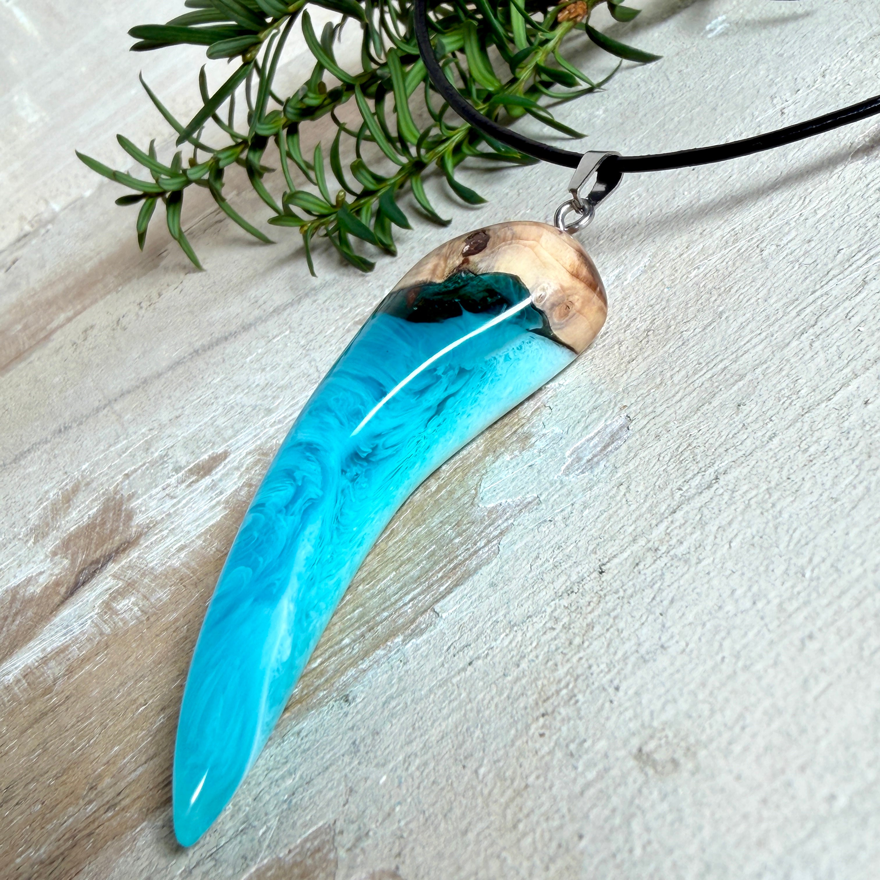 Handgefertigter Anhänger aus Edelholz und transparent-türkisblauem und weißen Epoxidharz, glatt poliert. Größe ca. 6,5 × 2,0 cm. Mit Edelstahlöse an schwarzer Lederkette (45 cm + 5 cm Edelstahl-Verlängerung, Karabiner). Moderner, unisex tragbarer Schmuck mit natürlicher Holzmaserung; kleine Lufteinschlüsse sind charakteristisch.