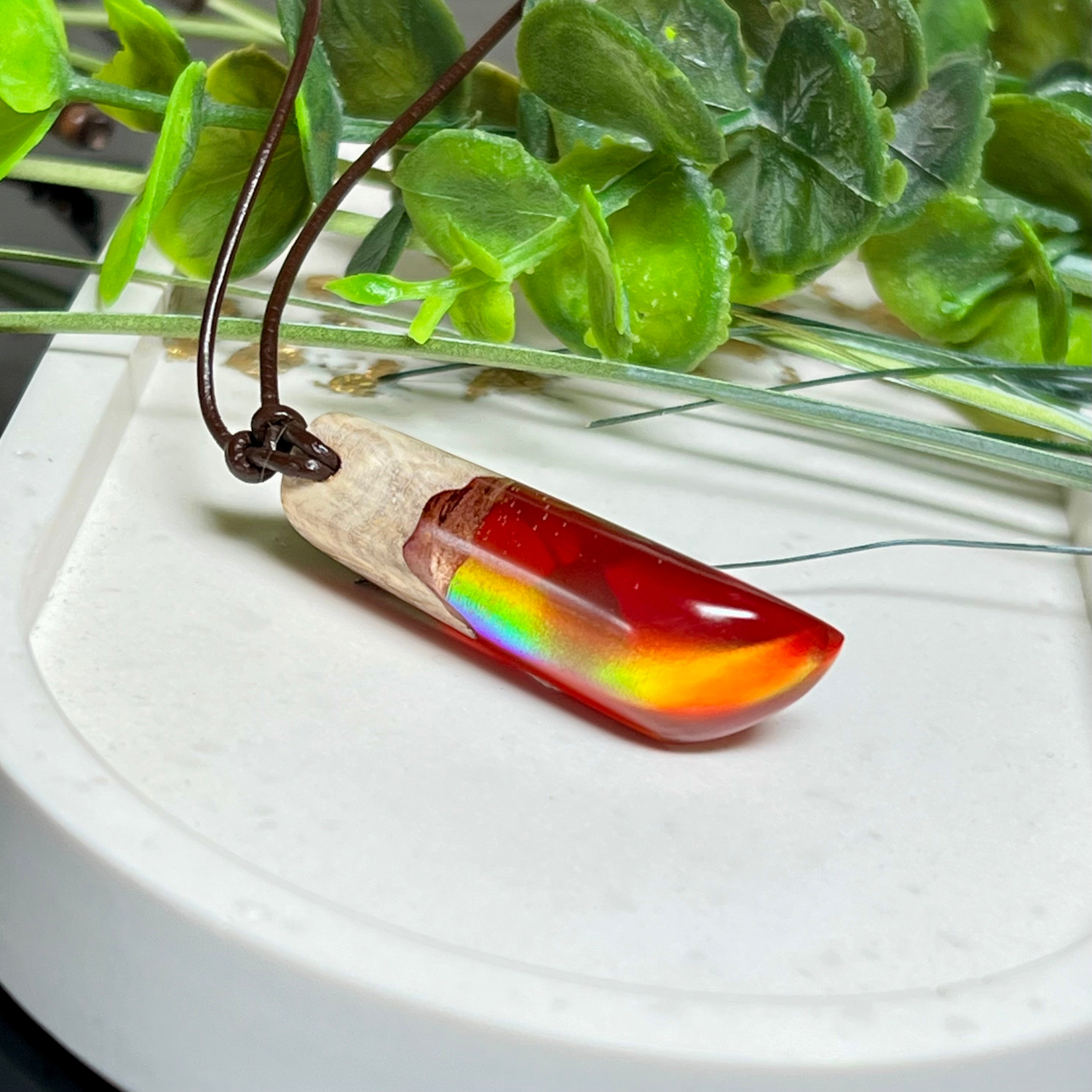 Handgefertigter Hei Toki Anhänger aus Goldfield-Maserholz und rubinrotem Labor-Opal, eingebettet in glänzendes Epoxidharz. Je nach Lichteinfall schimmert das Harz in warmen Farben mit Regenbogeneffekt. Der Anhänger ist ca. 4,3 × 1,5 cm groß und wird an einem verstellbaren Lederband mit Holzperlen getragen. Symbolischer Unikatschmuck – ideal als Geschenk für besondere Menschen.