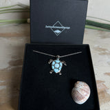 Feine Kette aus rhodiniertem 925 Silber mit Wasserschildkröten-Anhänger. Der ca. 25 x 18 mm große Anhänger zeigt eine Schildkröte, deren Panzer aus mehreren hellblauen Larimar-Inlays besteht. Seltener, kostbarer Larimar mit typischen wolkigen Strukturen, an einer ca. 45 cm langen, 1 mm starken, auf Platin rhodinierten Silberkette. Fotografiert in schwarzer Schmuckbox und vor maritimer Dekoration mit Muscheln.