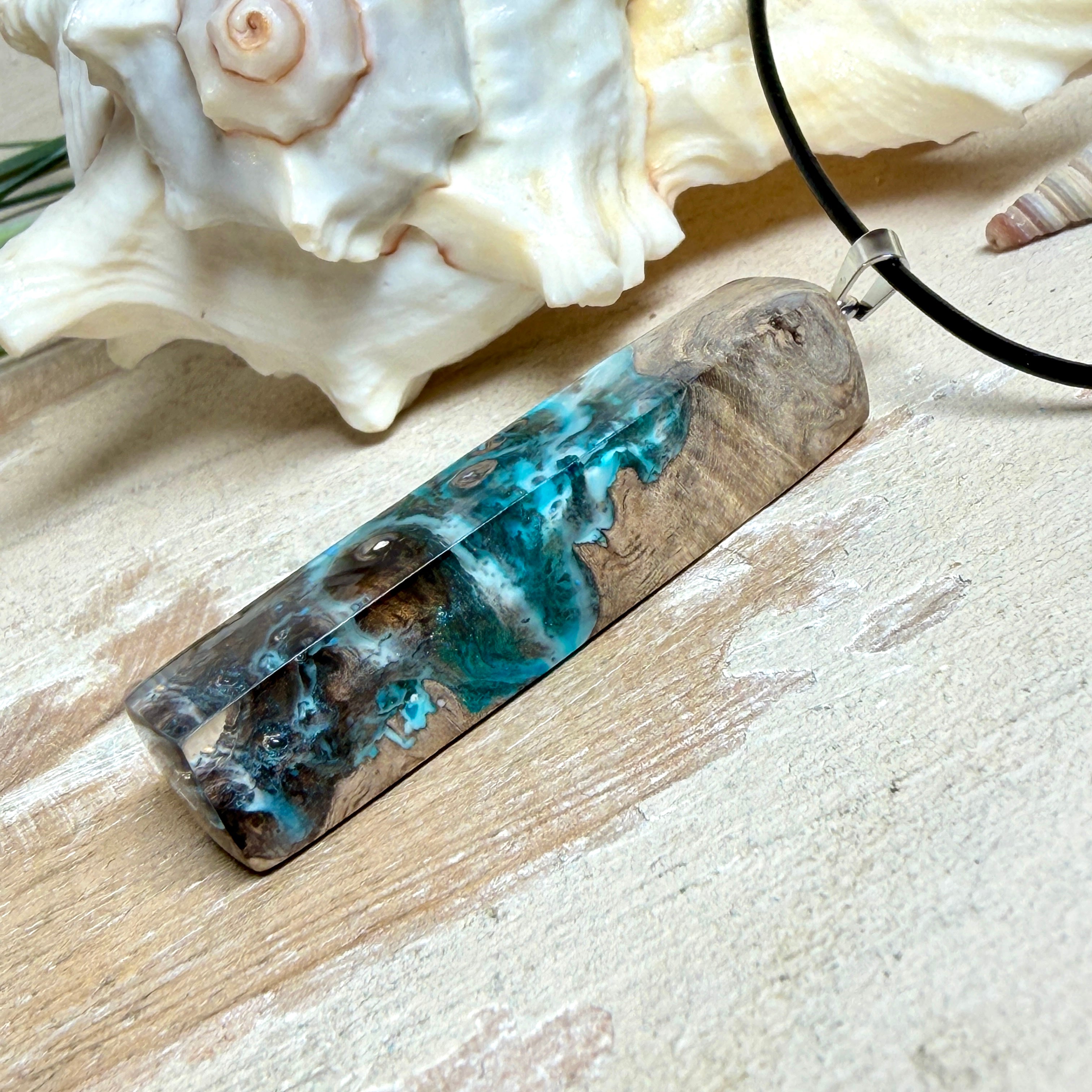 Rechteckiger Stabanhänger aus Maserholz und glasklarem Epoxidharz in tiefen Blau- und Türkistönen mit organischer Lagunen/Riff-Optik. Hochglanzpoliert, ca. 5,3 × 1,2 cm. Der Anhänger hängt an einer schwarzen Lederkette mit Edelstahl-Karabiner und 5-cm-Verlängerung (45 + 5 cm); die Anhängerschlaufe ist ebenfalls aus Edelstahl. Handgefertigtes Unikat, Lieferung in edler Schmuckbox – maritimer Look.