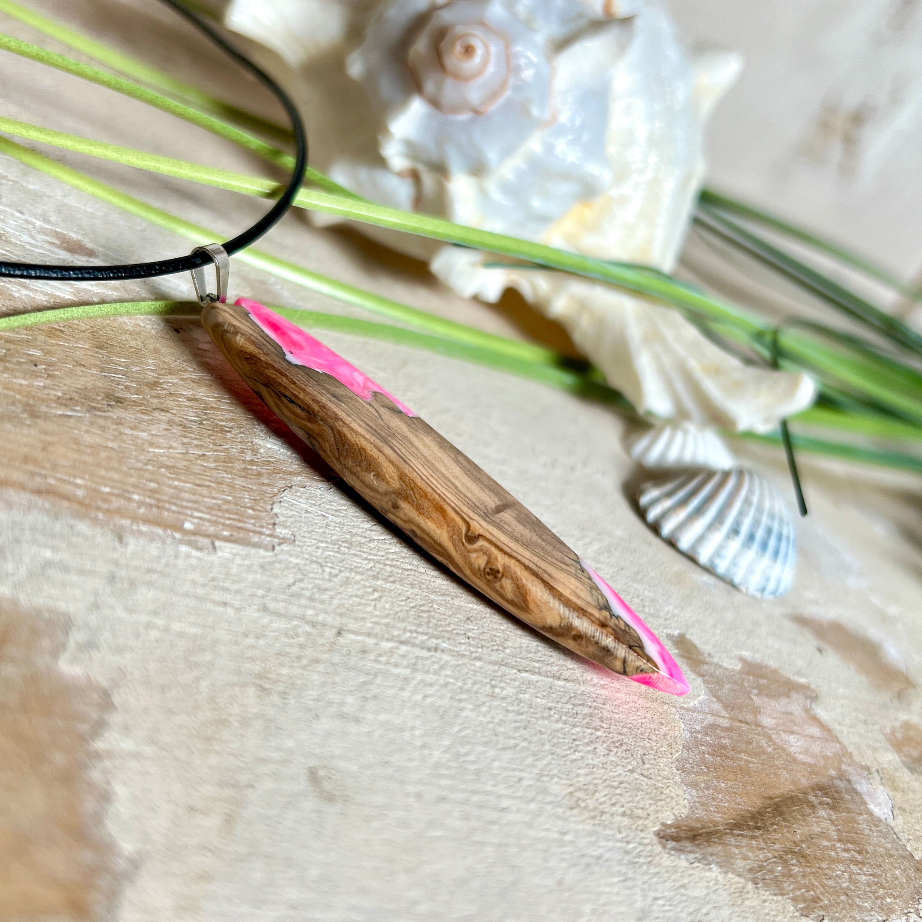 Handgefertigter Surfbrett-Anhänger „Pink Glow“ aus edlem Olivenholz und fluoreszierendem Epoxidharz in Pink & Weiß. Fluoreszierend bei Schwarzlicht, mit Edelstahl-Anhängerschlaufe und schwarzer Lederkette (45 cm + 5 cm Verlängerung). Ein maritimes Unikat – perfekt als Geschenk für Surfer und Meeresliebhaber.