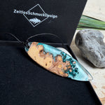Handgefertigter, länglicher Surfbrett-Anhänger aus naturbelassenem Maserholz und transparentem Harz in Lagunen-Türkis, an schwarzem Lederband mit Knoten – maritimer Unikatschmuck für Damen und Herren, dekorativ mit Stein und Muschel inszeniert.