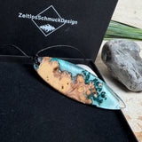 Handgefertigter, länglicher Surfbrett-Anhänger aus naturbelassenem Maserholz und transparentem Harz in Lagunen-Türkis, an schwarzem Lederband mit Knoten – maritimer Unikatschmuck für Damen und Herren, dekorativ mit Stein und Muschel inszeniert.