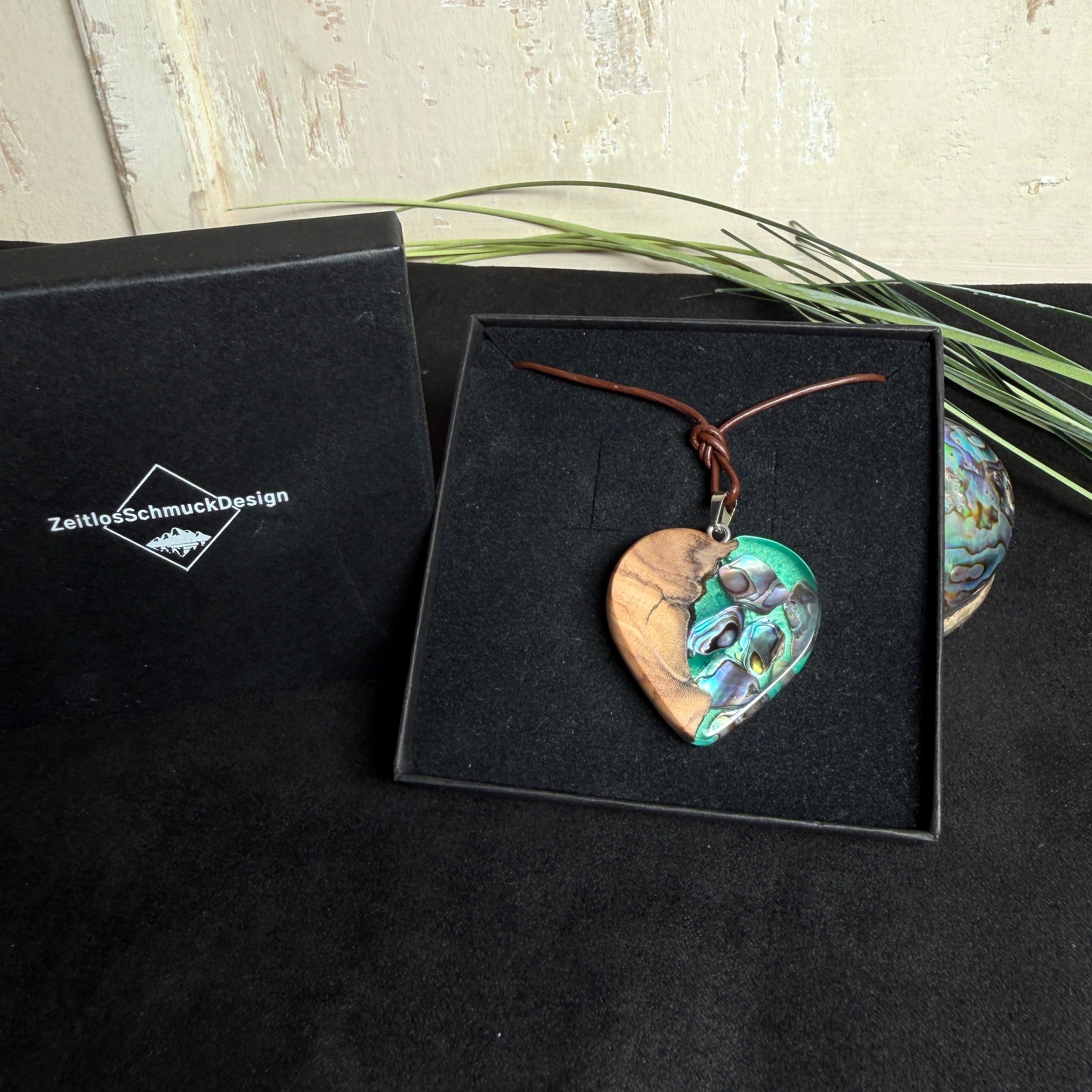 Herzförmiger Anhänger aus Olivenholz mit aquamarinfarbenem Harz und eingelegten Paua-Muschelstücken (Abalone), irisierender Blau-Grün-Schimmer. Der Anhänger hängt an einem braunen, verstellbaren Lederband mit Schiebeknoten; Aufhängung aus Edelstahl. Produktfoto vor maritimer Deko/Abalone-Schale.