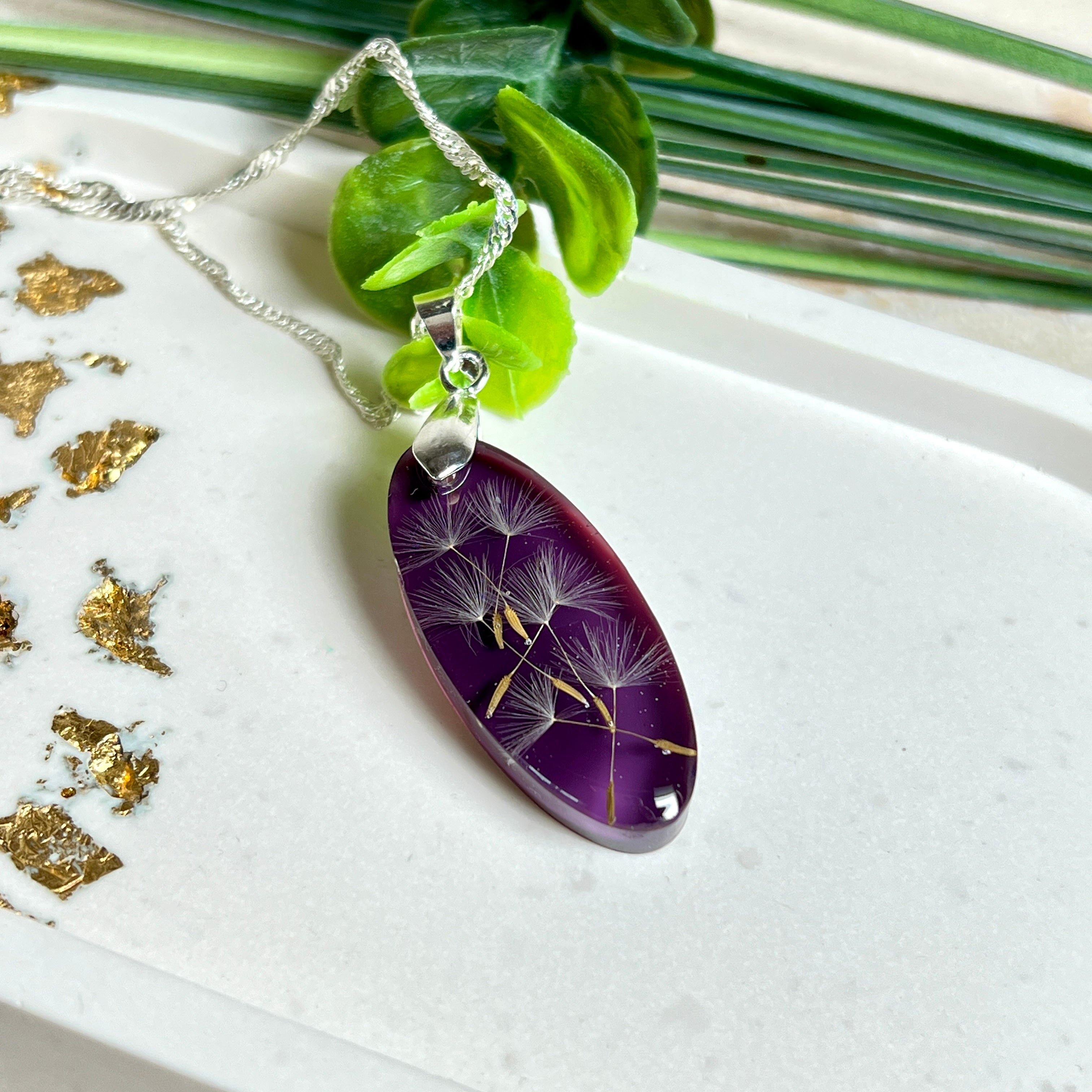 Ovaler Anhänger mit echter Pusteblume in Harz, Hintergrund Brombeerfarben. Größe 4 × 1,8 cm, versilberte Kette 50 cm mit Karabinerverschluss. Handgefertigtes Unikat.