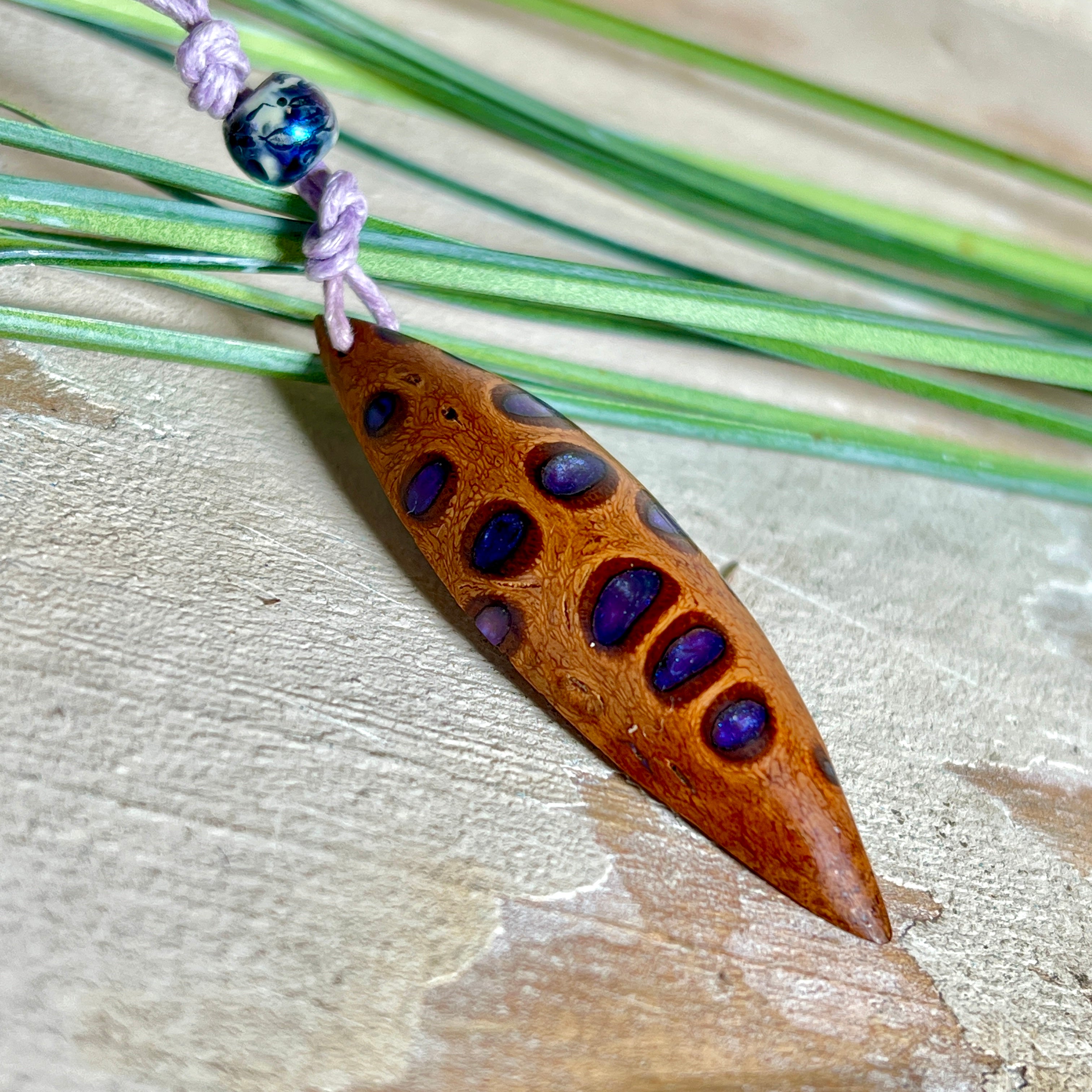 Handgefertigter Anhänger aus einem australischen Banksiazapfen mit eingelassenem metallic-violettem Harz. Der ovale, tropfenförmige Anhänger ist ca. 5,5 cm lang und wird mit einem verstellbaren Baumwollband geliefert. Jedes Stück zeigt die einzigartige natürliche Struktur des Zapfens und schimmert in satten Lilatönen – ein stilvoller Schmuck mit organischem Charakter.