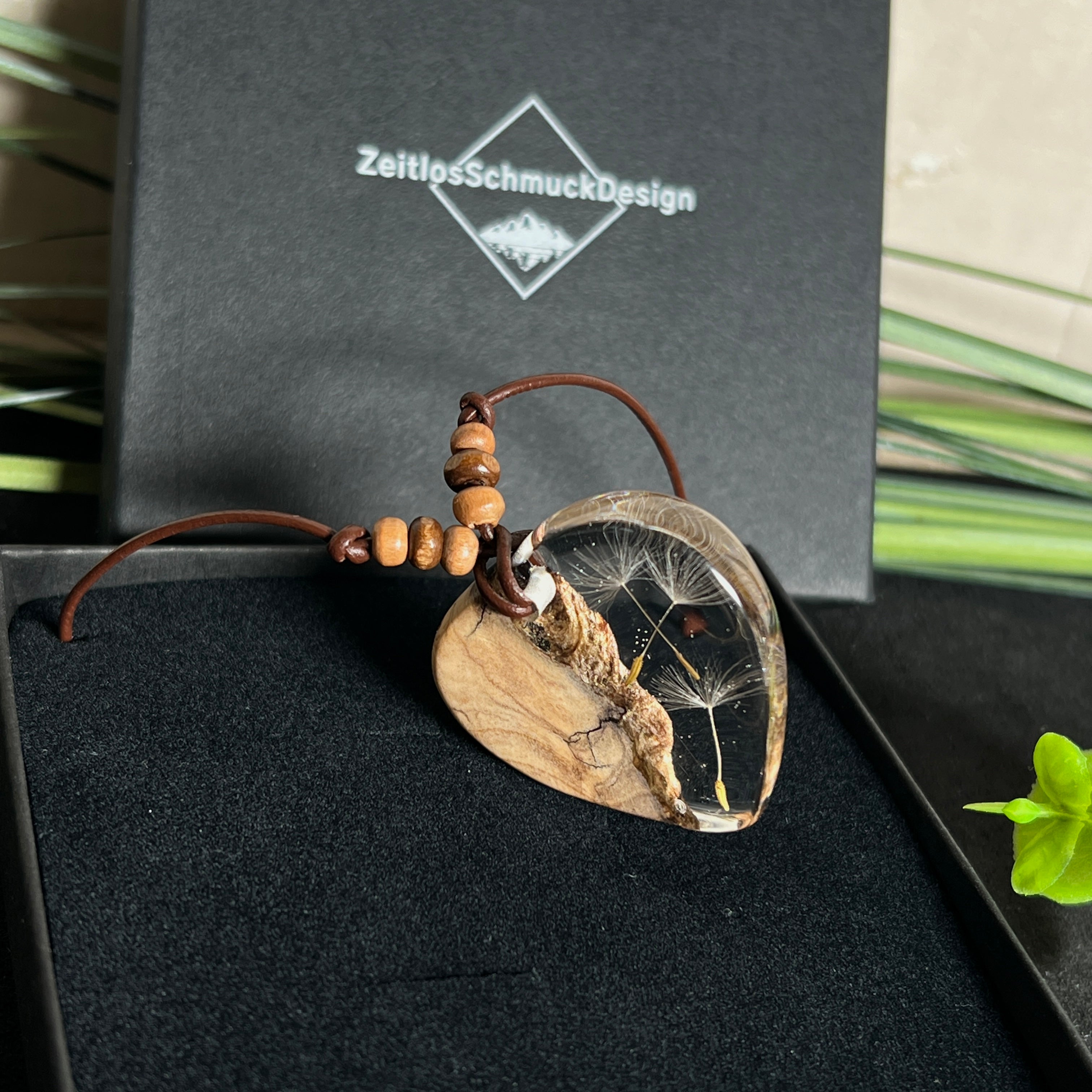 Handgefertigter Herzanhänger aus Olivenholz und klarem Harz mit echten Pusteblumen. Der Anhänger misst ca. 3,5 cm, das Lederband ist verstellbar von 23–43 cm und mit kleinen Holzperlen verziert. Ein natürliches Unikat, Symbol für Wünsche und Glück – sorgfältig gegossen, gesägt, geschliffen und poliert.