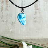 Larimar Freeform-Anhänger auf schwarzer Lederkette. Beidseitig tragbarer Trommelstein, ca. 2 x 1,8 cm groß, mit Silberöse. Der seltene, kostbare Larimar zeigt lebendige Blau- und Weißnuancen wie karibisches Lagunenwasser. Lederkette 45 cm mit Edelstahl-Karabiner und 5 cm Edelstahl-Verlängerung, fotografiert vor einer großen Muschel im maritimen Stil.