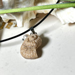 Handgefertigter Anhänger in Ammonit-Form aus echtem Meersand und Harz, kombiniert mit einer weißen Süßwasserperle. Der maritime Anhänger hängt an einer schwarzen Lederkette mit Edelstahlöse und Karabinerverschluss. Im Hintergrund liegt eine weiße Muschel, die das natürliche, strandinspirierte Design des Schmuckstücks unterstreicht. Ideal als besonderes Geschenk oder Erinnerung an den Sommer.