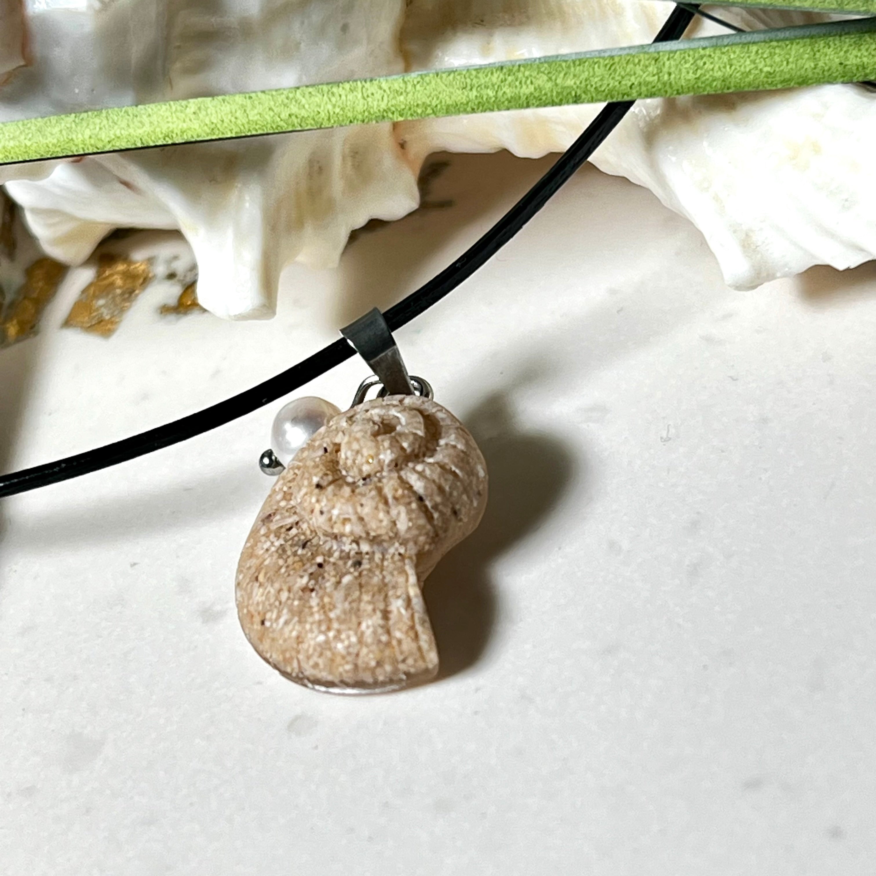 Handgefertigter Anhänger in Ammonit-Form aus echtem Meersand und Harz, kombiniert mit einer weißen Süßwasserperle. Der maritime Anhänger hängt an einer schwarzen Lederkette mit Edelstahlöse und Karabinerverschluss. Im Hintergrund liegt eine weiße Muschel, die das natürliche, strandinspirierte Design des Schmuckstücks unterstreicht. Ideal als besonderes Geschenk oder Erinnerung an den Sommer.