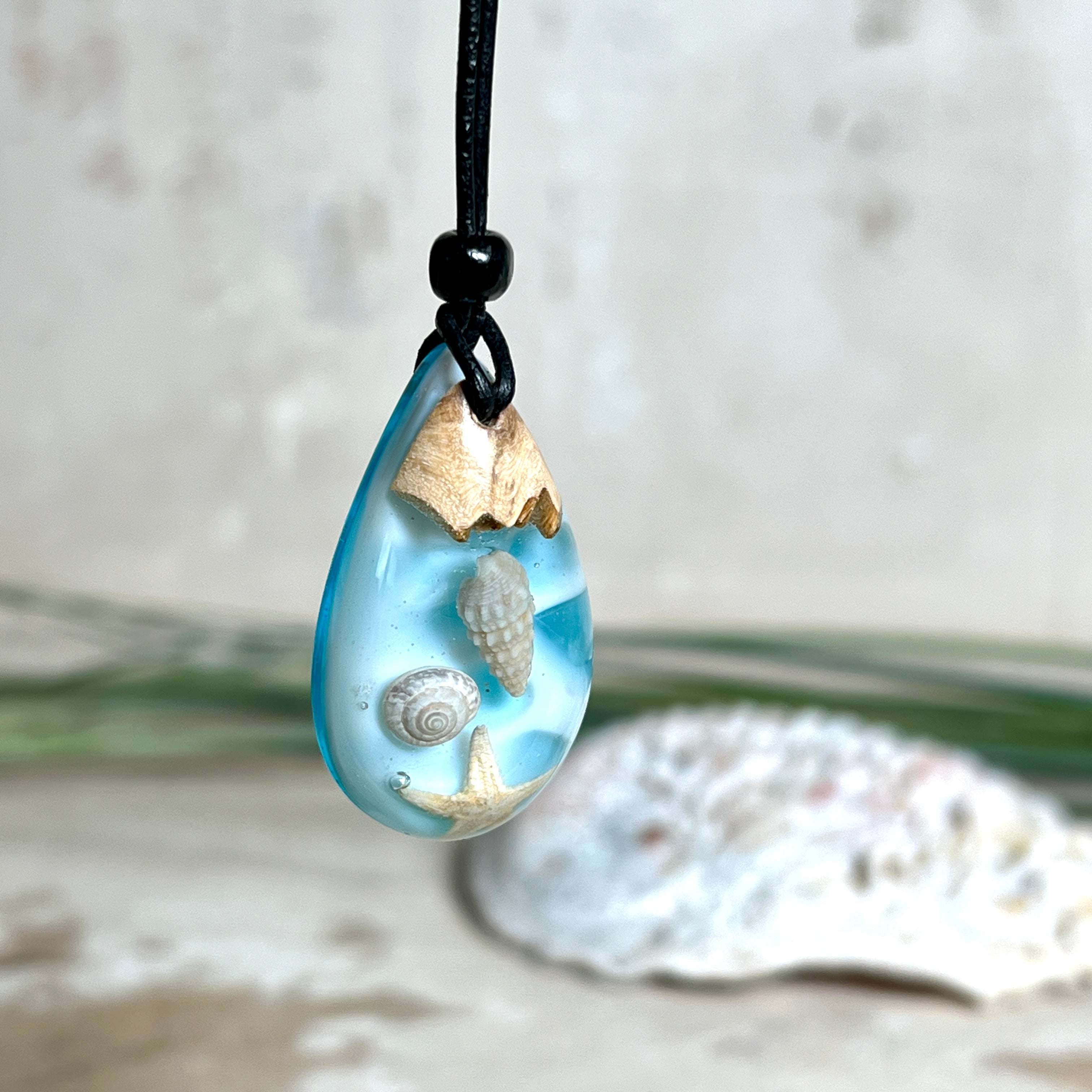 Handgefertigter Tropfenanhänger aus Maserholz und klarem Epoxidharz in Aquatönen. Im Harz sind echte Muscheln und ein kleiner Seestern eingebettet. Durch die besondere Mehrschicht-Gießtechnik wirkt es, als würden die Muscheln im Wasser schweben. Größe ca. 3,8 × 2 cm. Verstellbares Lederband (24–44 cm) mit Holzperlen. Maritimes Unikat.