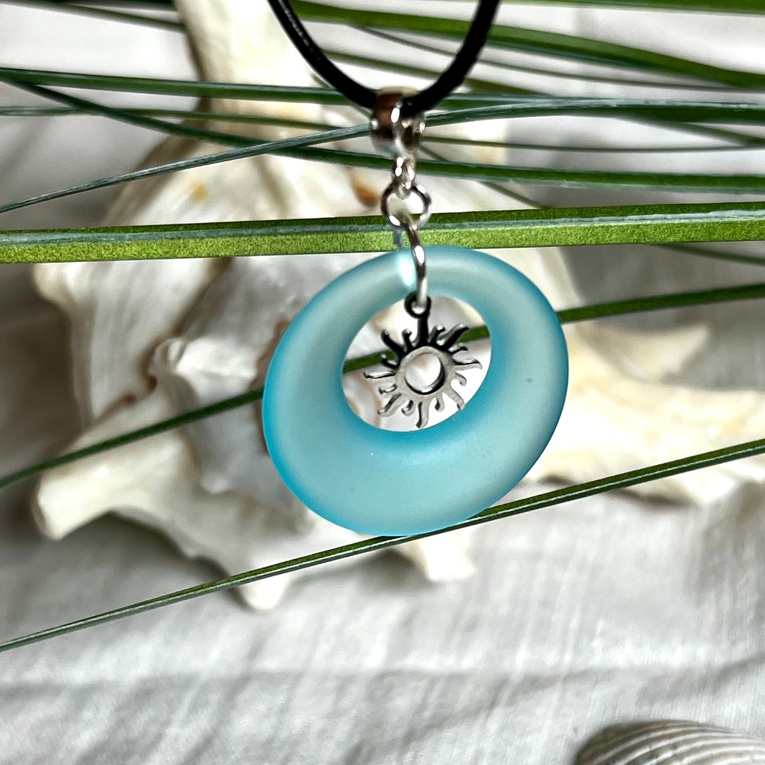 Handgefertigte Halskette mit Donut-Anhänger aus türkisfarbenem Seeglas, kombiniert mit einem filigranen Sonnen-Charm aus Edelstahl. Die Kette besteht aus einer schwarzen Lederschnur (1,5 mm) mit Karabiner und Verlängerungskette aus Edelstahl. Maritimes Schmuckstück für Meerliebhaberinnen und Sommerfreunde. Unikat mit stilvoller Ausstrahlung.
