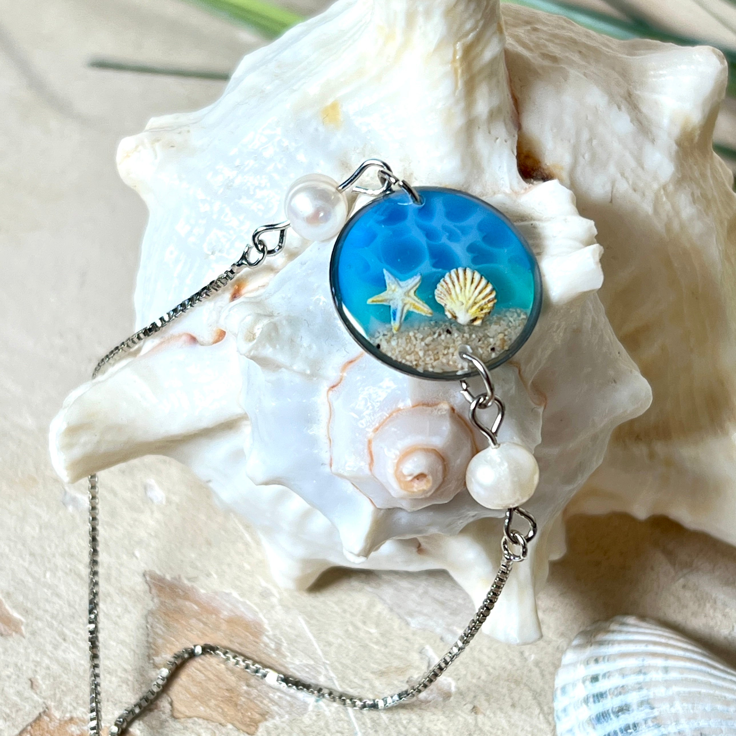Elegantes Edelstahl-Armband mit rundem Resin-Anhänger in Meeresblau. Im Anhänger liegt echter Meersand als Ufersaum; darüber Sticker-Motive Seestern & Muschel. Zwei feine Süßwasserperlen verzieren das Armband. Verstellbarer Schiebeverschluss . Handgefertigtes Unikat – maritimer Schmuck, der an Wellenrauschen und Strandspaziergänge erinnert.