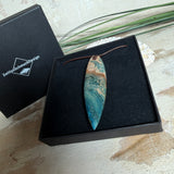 Unikat Anhänger aus stabilisiertem Holz mit türkisblauem Harzverlauf, schlanke Form, poliert. Verstellbares braunes Lederband 22–46 cm mit Schiebeknoten. Anhänger ca. 6,6 × 2 cm. Tragefoto am Hals und Produktfotos mit maritimer Deko