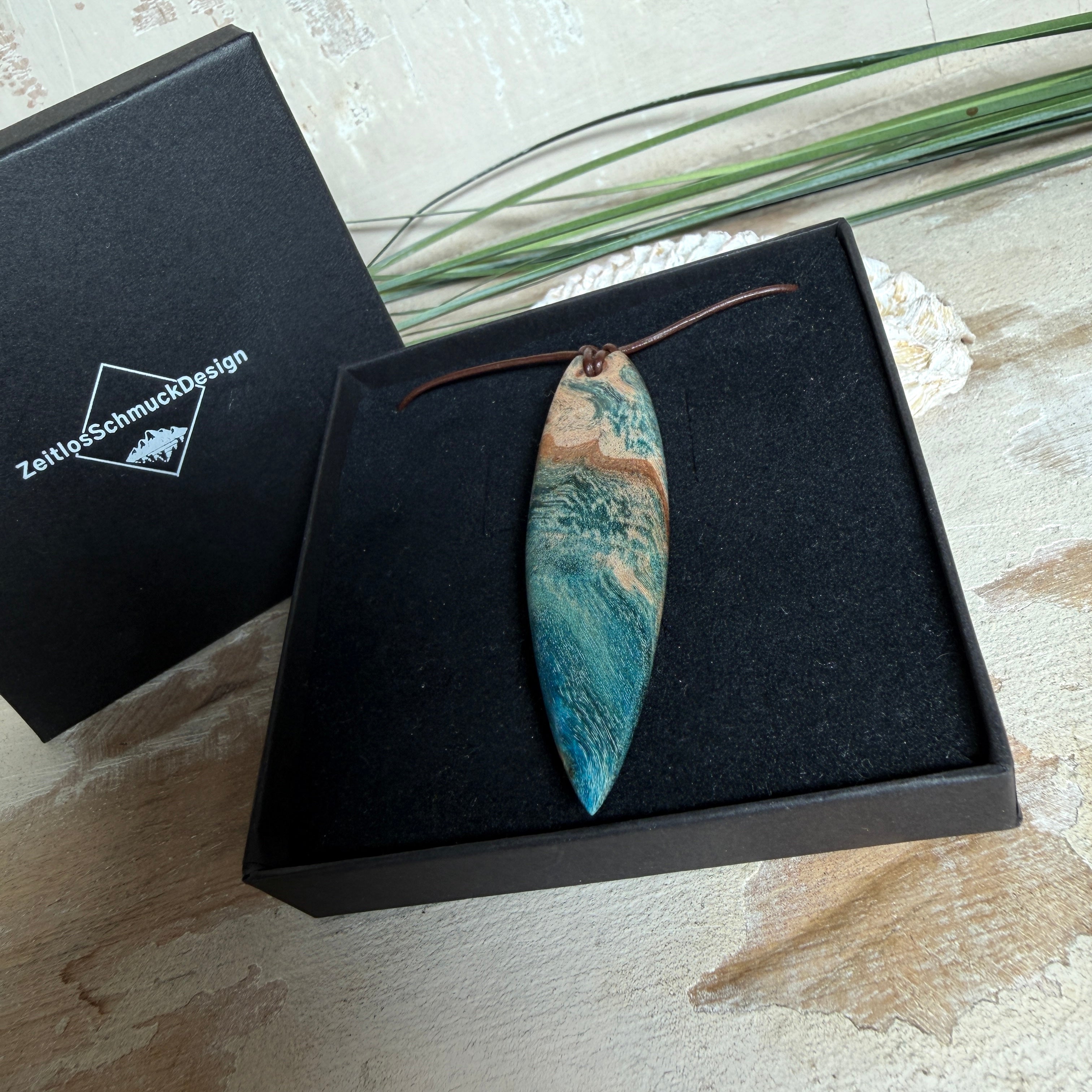 Unikat Anhänger aus stabilisiertem Holz mit türkisblauem Harzverlauf, schlanke Form, poliert. Verstellbares braunes Lederband 22–46 cm mit Schiebeknoten. Anhänger ca. 6,6 × 2 cm. Tragefoto am Hals und Produktfotos mit maritimer Deko
