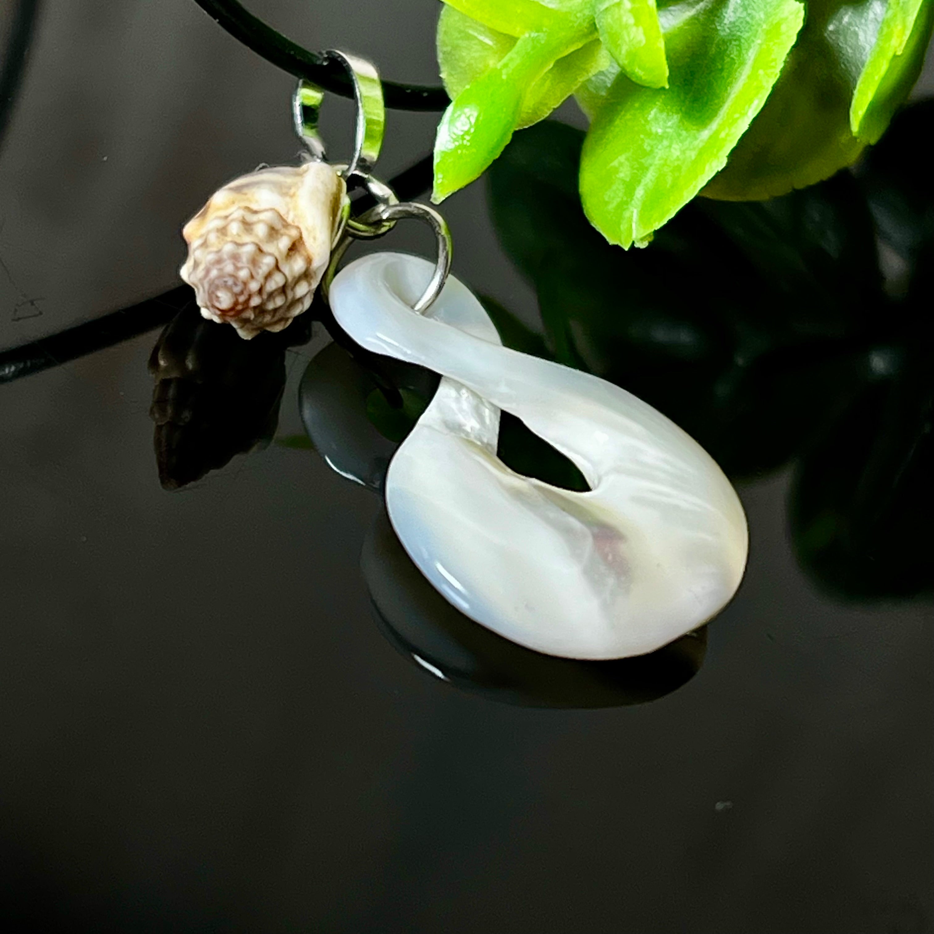 Detailaufnahme eines handgefertigten Anhängers aus echtem Perlmutt in Schlaufenform, kombiniert mit einer kleinen echten Muschel als Charm-Anhänger. Der Schmuck hängt an einer feinen schwarzen Lederschnur mit Edelstahlverschluss und symbolisiert Harmonie und Unendlichkeit. Ideal für Meeresliebhaberinnen und Fans von Naturmaterialien.