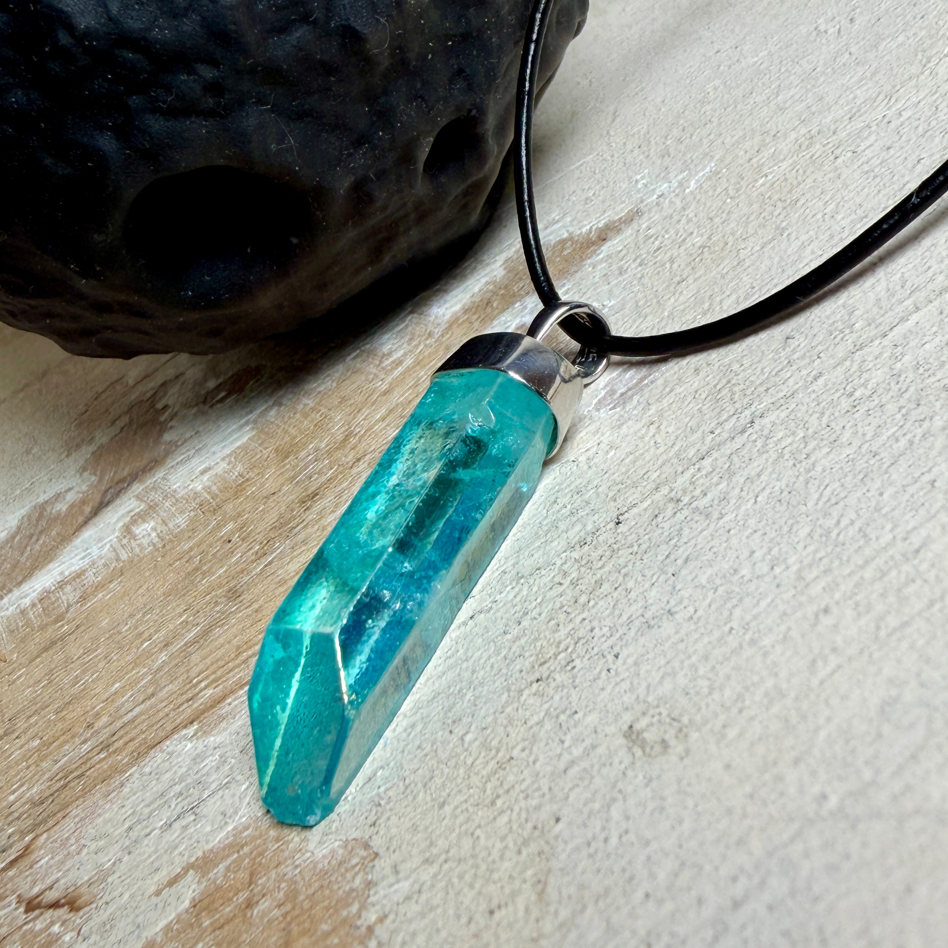 Nahaufnahme einer handgefertigten Halskette mit Aqua Aura Rohkristall: naturbelassener, sechskantiger Bergkristall mit goldbedampfter Aqua-Beschichtung in Türkisblau, gefasst in 925er Silber und getragen an einer schwarzen Lederkette mit Edelstahl-Karabiner und Verlängerungskettchen.