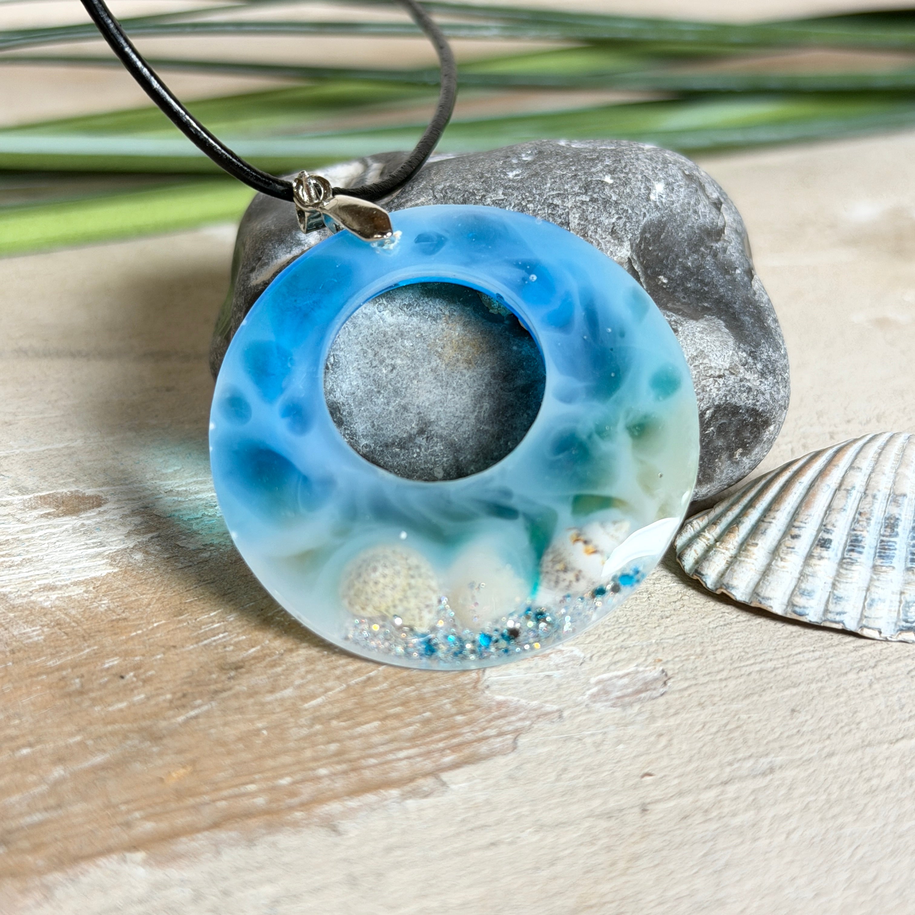 Runder Harzanhänger „Ocean Circle“ mit 4 cm Durchmesser in Blau-Weiß-Verlauf, in dem kleine Muscheln und feiner Glitzer wie ein Mini-Strand eingefangen sind. Der Anhänger hat eine Edelstahl-Schlaufe und hängt an einer schwarzen Lederkette mit Edelstahl-Karabiner und Verlängerung. Maritimer, handgefertigter Unikat-Schmuck im Lagunen-Look für Meeresliebhaberinnen.
