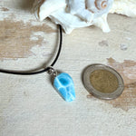 Larimar Freeform-Anhänger auf schwarzer Lederkette. Beidseitig tragbarer Trommelstein, ca. 2 x 1,8 cm groß, mit Silberöse. Der seltene, kostbare Larimar zeigt lebendige Blau- und Weißnuancen wie karibisches Lagunenwasser. Lederkette 45 cm mit Edelstahl-Karabiner und 5 cm Edelstahl-Verlängerung, fotografiert vor einer großen Muschel im maritimen Stil.