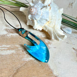 Handgefertigter Haken-Anhänger im Hei-Matau-Stil aus Maserholz und klarem Harz mit Blau-, Türkis- und Weißverlauf, hochglanzpoliert. Oben sichtbares Holz im Harzbereich, getragen an verstellbarem schwarzem Lederband mit Schiebeknoten und Zierperlen. Anhänger ca. 7 × 3 cm, fotografiert mit Muscheln im Hintergrund.