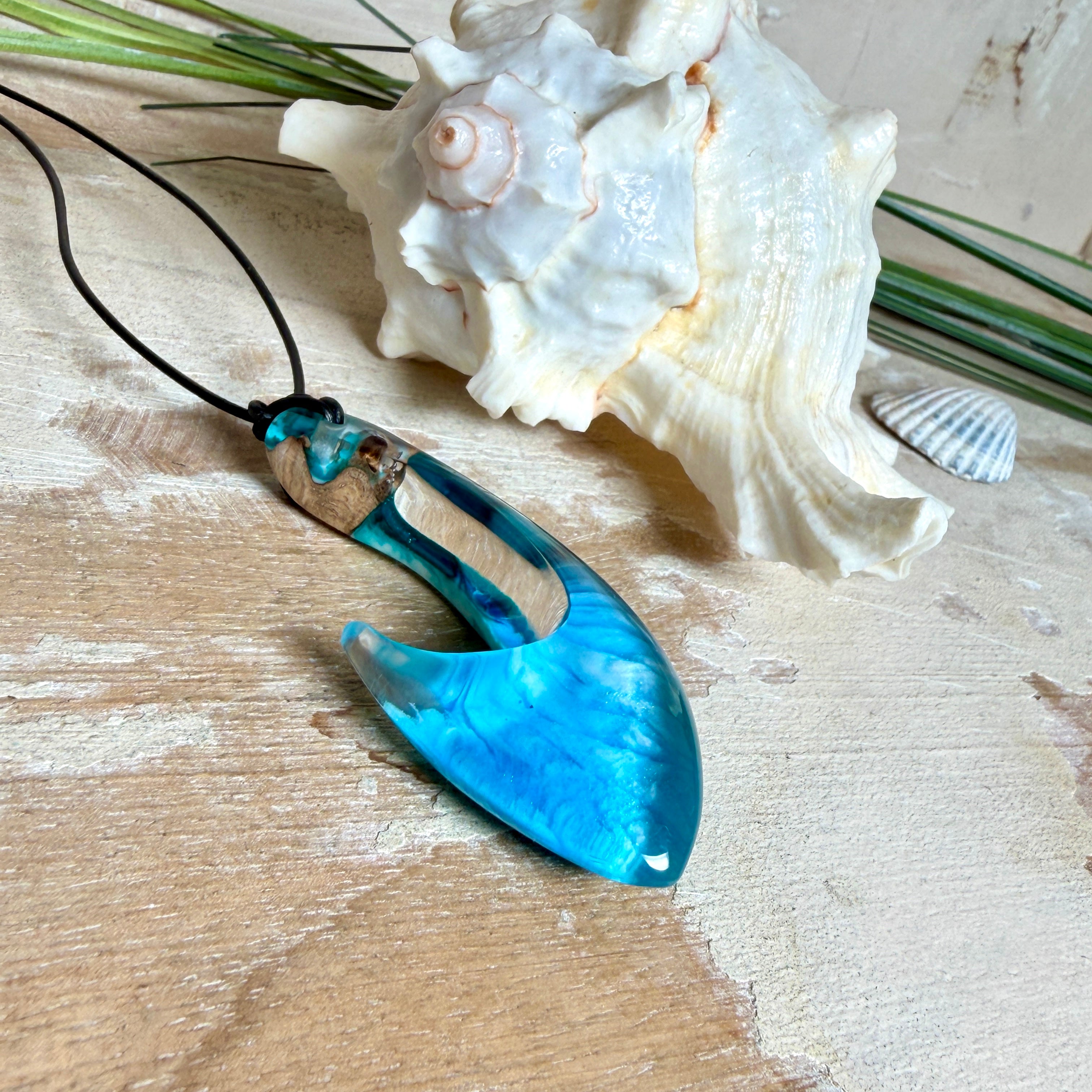 Handgefertigter Haken-Anhänger im Hei-Matau-Stil aus Maserholz und klarem Harz mit Blau-, Türkis- und Weißverlauf, hochglanzpoliert. Oben sichtbares Holz im Harzbereich, getragen an verstellbarem schwarzem Lederband mit Schiebeknoten und Zierperlen. Anhänger ca. 7 × 3 cm, fotografiert mit Muscheln im Hintergrund.