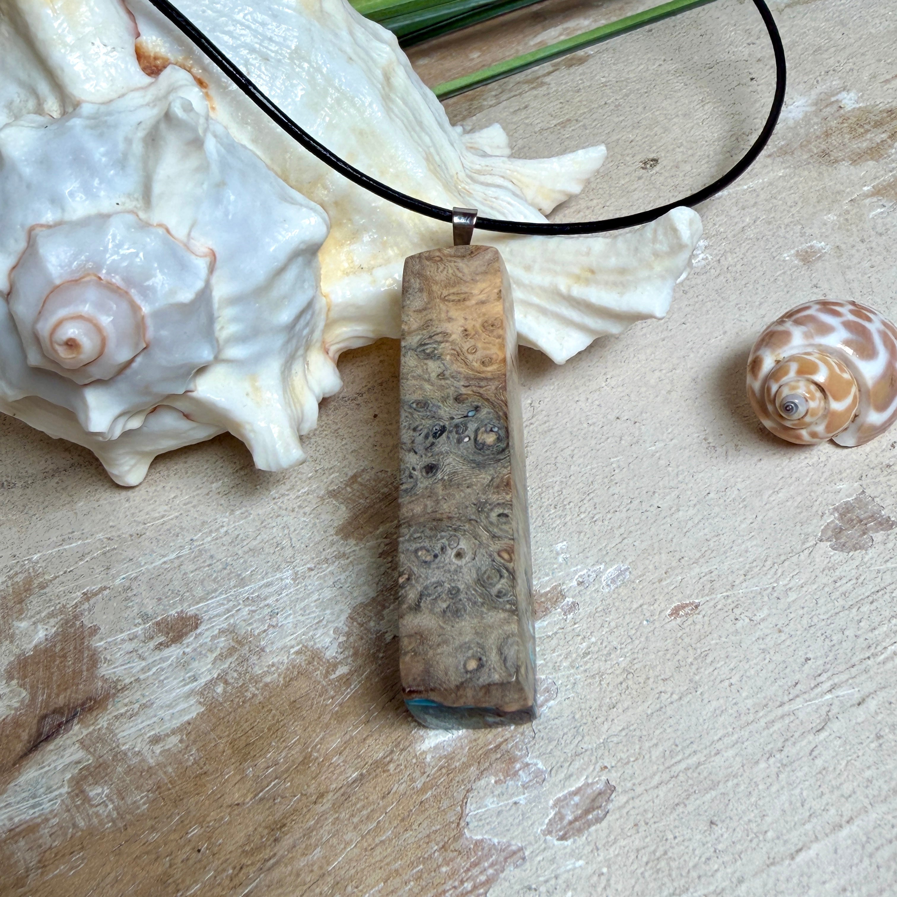 Rechteckiger Stabanhänger aus Maserholz und glasklarem Epoxidharz in tiefen Blau- und Türkistönen mit organischer Lagunen/Riff-Optik. Hochglanzpoliert, ca. 5,3 × 1,2 cm. Der Anhänger hängt an einer schwarzen Lederkette mit Edelstahl-Karabiner und 5-cm-Verlängerung (45 + 5 cm); die Anhängerschlaufe ist ebenfalls aus Edelstahl. Handgefertigtes Unikat, Lieferung in edler Schmuckbox – maritimer Look.