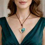 Herzförmiger Anhänger aus Olivenholz mit aquamarinfarbenem Harz und eingelegten Paua-Muschelstücken (Abalone), irisierender Blau-Grün-Schimmer. Der Anhänger hängt an einem braunen, verstellbaren Lederband mit Schiebeknoten; Aufhängung aus Edelstahl. Produktfoto vor maritimer Deko/Abalone-Schale.