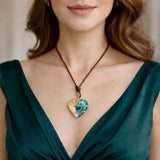 Herzförmiger Anhänger aus Olivenholz mit aquamarinfarbenem Harz und eingelegten Paua-Muschelstücken (Abalone), irisierender Blau-Grün-Schimmer. Der Anhänger hängt an einem braunen, verstellbaren Lederband mit Schiebeknoten; Aufhängung aus Edelstahl. Produktfoto vor maritimer Deko/Abalone-Schale.