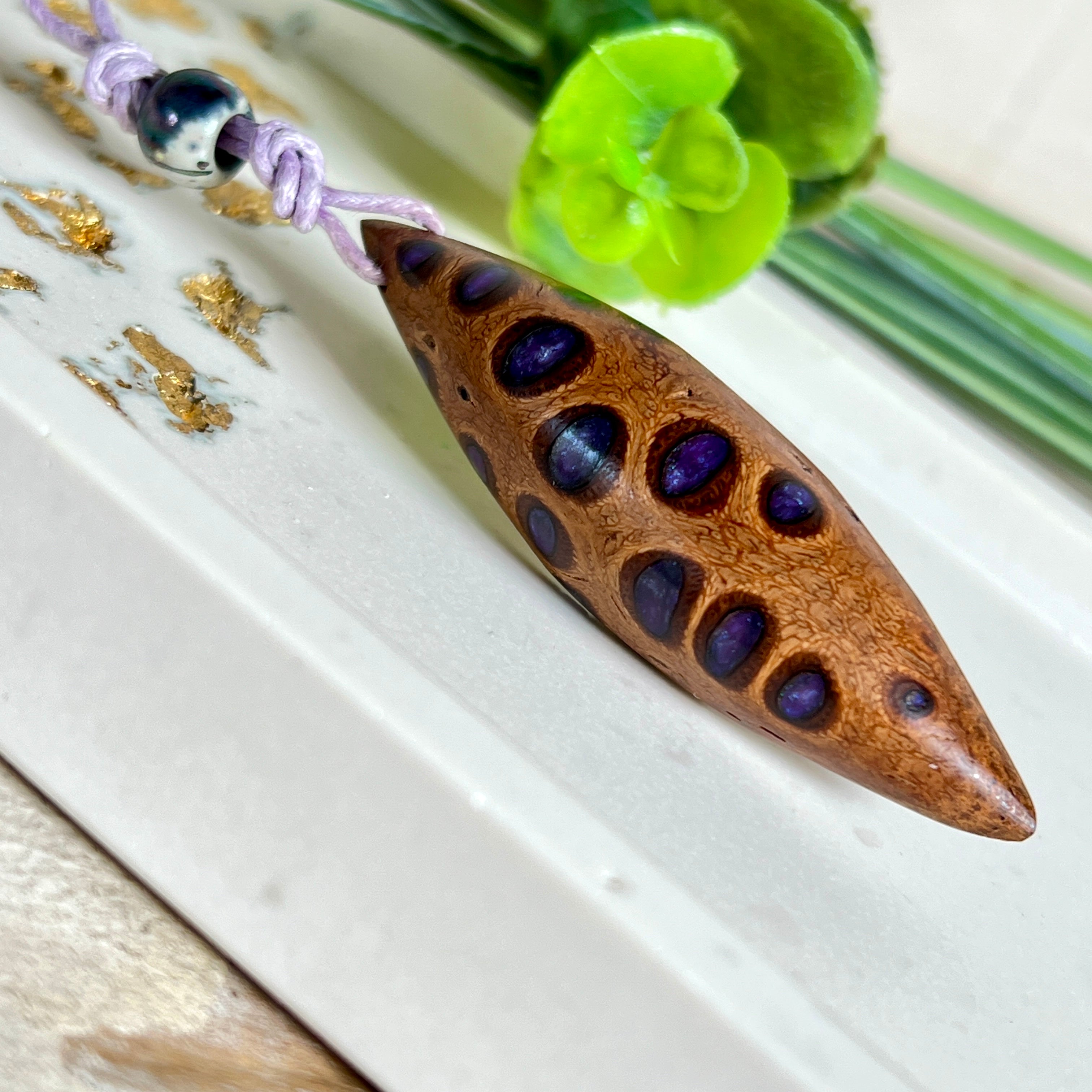 Handgefertigter Anhänger aus einem australischen Banksiazapfen mit eingelassenem metallic-violettem Harz. Der ovale, tropfenförmige Anhänger ist ca. 5,5 cm lang und wird mit einem verstellbaren Baumwollband geliefert. Jedes Stück zeigt die einzigartige natürliche Struktur des Zapfens und schimmert in satten Lilatönen – ein stilvoller Schmuck mit organischem Charakter.