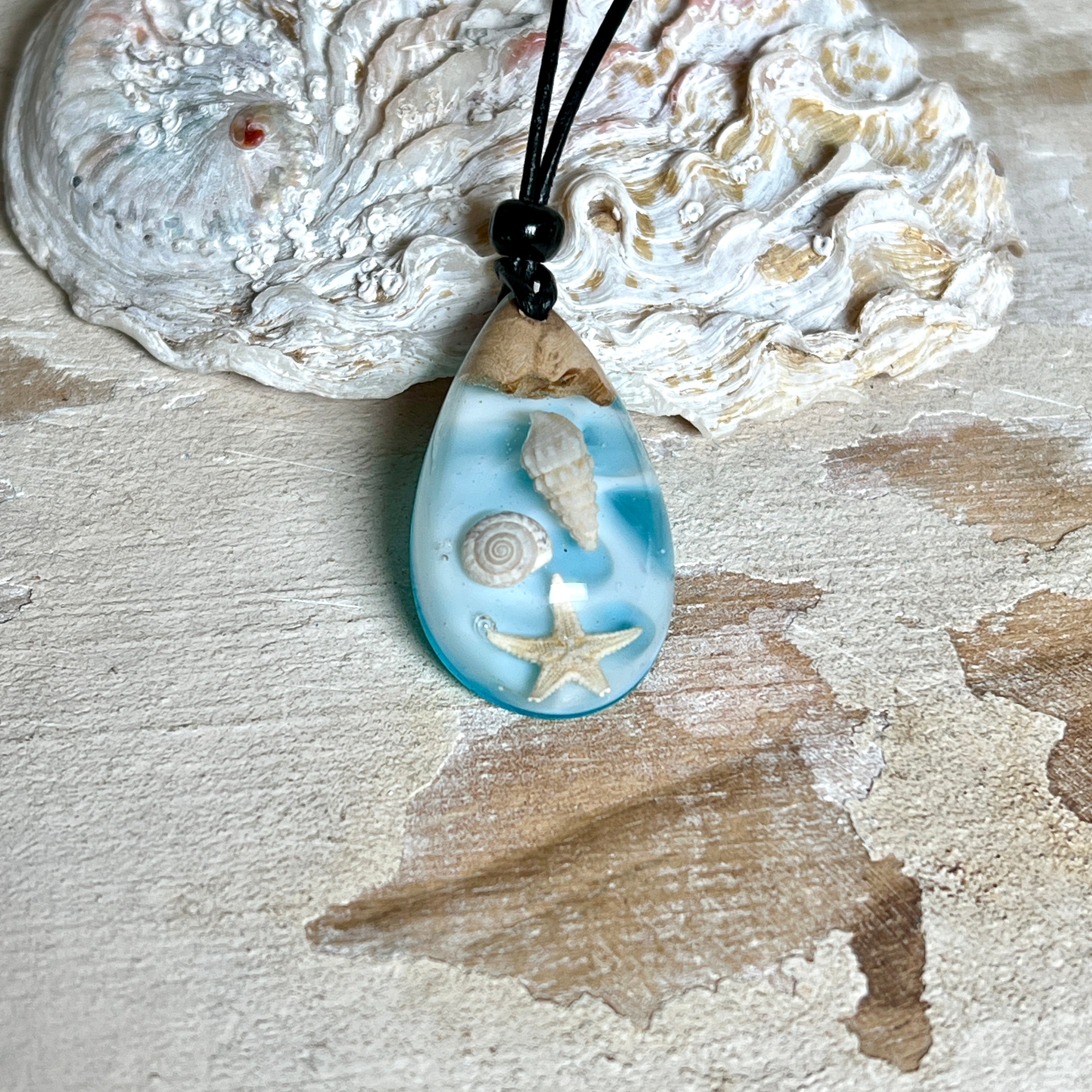 Handgefertigter Tropfenanhänger aus Maserholz und klarem Epoxidharz in Aquatönen. Im Harz sind echte Muscheln und ein kleiner Seestern eingebettet. Durch die besondere Mehrschicht-Gießtechnik wirkt es, als würden die Muscheln im Wasser schweben. Größe ca. 3,8 × 2 cm. Verstellbares Lederband (24–44 cm) mit Holzperlen. Maritimes Unikat.
