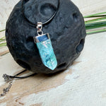 Nahaufnahme einer handgefertigten Halskette mit Aqua Aura Rohkristall: naturbelassener, sechskantiger Bergkristall mit goldbedampfter Aqua-Beschichtung in Türkisblau, gefasst in 925er Silber und getragen an einer schwarzen Lederkette mit Edelstahl-Karabiner und Verlängerungskettchen.