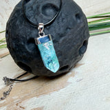 Nahaufnahme einer handgefertigten Halskette mit Aqua Aura Rohkristall: naturbelassener, sechskantiger Bergkristall mit goldbedampfter Aqua-Beschichtung in Türkisblau, gefasst in 925er Silber und getragen an einer schwarzen Lederkette mit Edelstahl-Karabiner und Verlängerungskettchen.