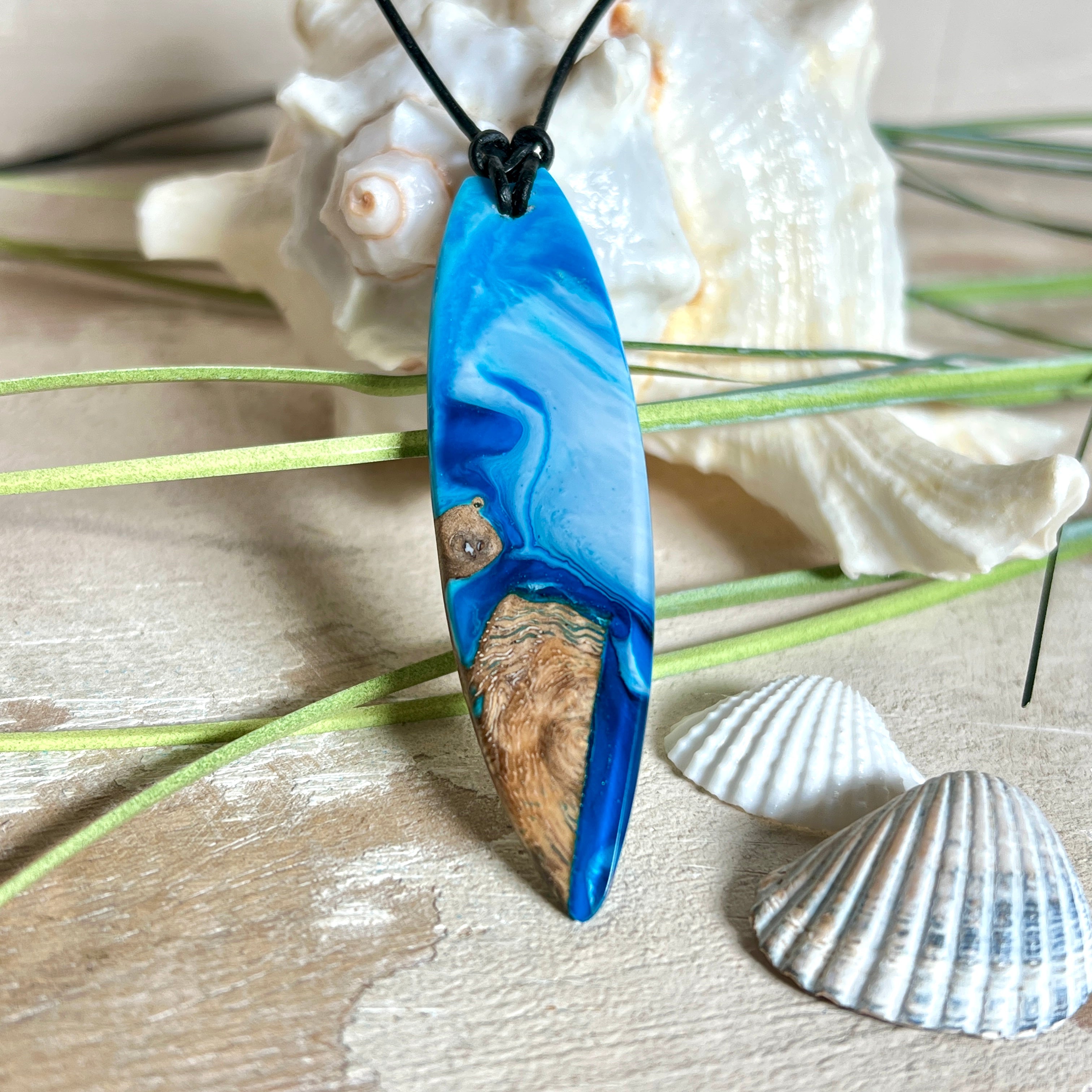 Handgefertigter Surfbrett-Anhänger „Ocean Glide“ aus Maserholz und Epoxidharz in verschiedenen Blau-, und Weißtönen. Der ca. 6,5 × 2 cm große Anhänger wird an einem verstellbaren Lederband (26–46 cm) mit Holzperlen geliefert. Ein maritimes Unikat mit natürlicher Holzmaserung, das die Schönheit des Meeres einfängt.