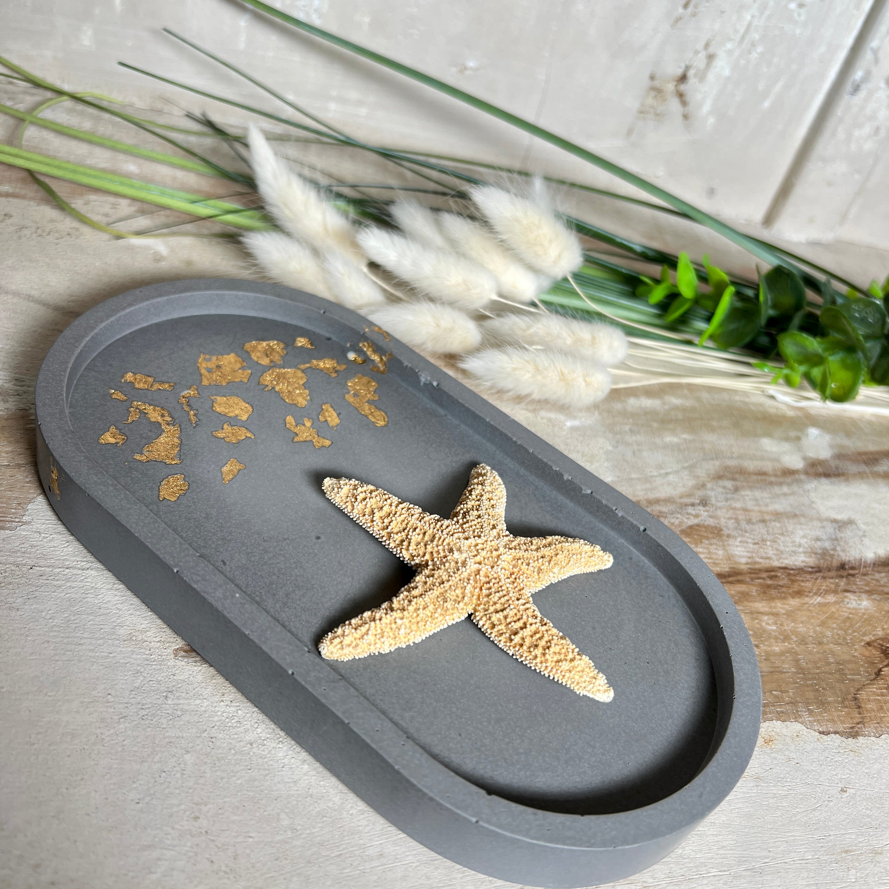 Handgefertigtes ovales Schmucktablett aus grauen  Jesmonite mit schimmernden Blattgold-Details. Die Unterseite ist mit vier Korkpunkten versehen und schützt empfindliche Oberflächen. Ideal zur stilvollen Aufbewahrung von Schmuck, Schlüsseln, Brille oder kleinen Dekoartikeln. Jedes Tablett ist ein Unikat mit individueller Maserung und goldenen Akzenten – Lieferung ohne Deko. Die 